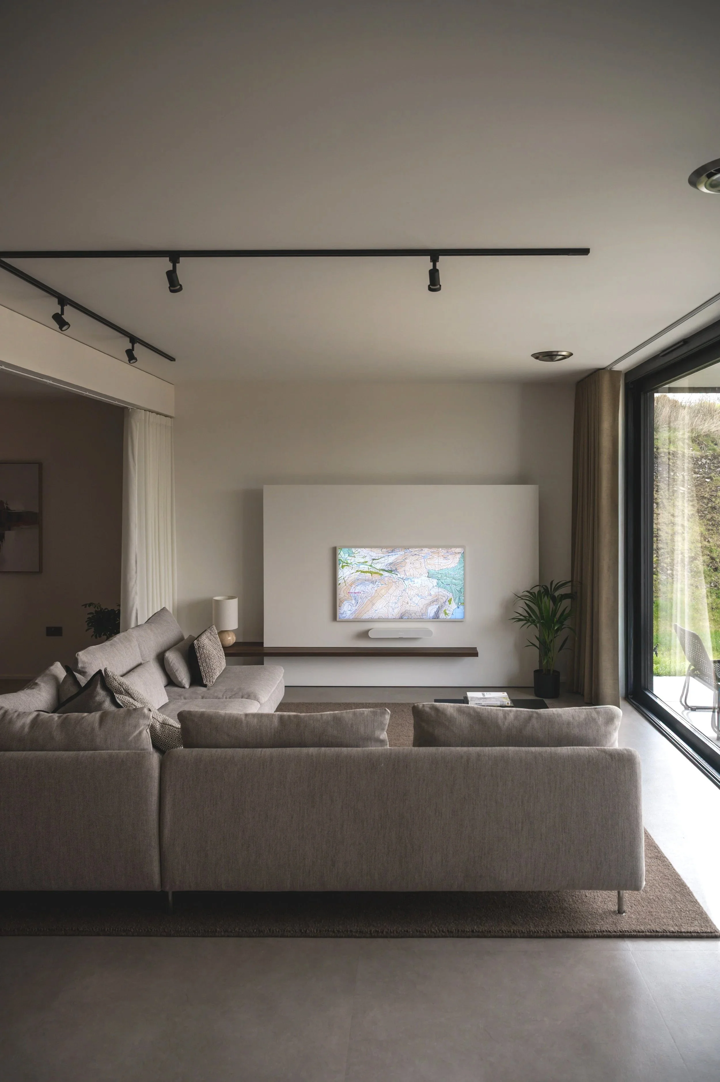 A modern living room with beige sofas, a wall-mounted television, a potted plant, and large windows with curtains letting in natural light.