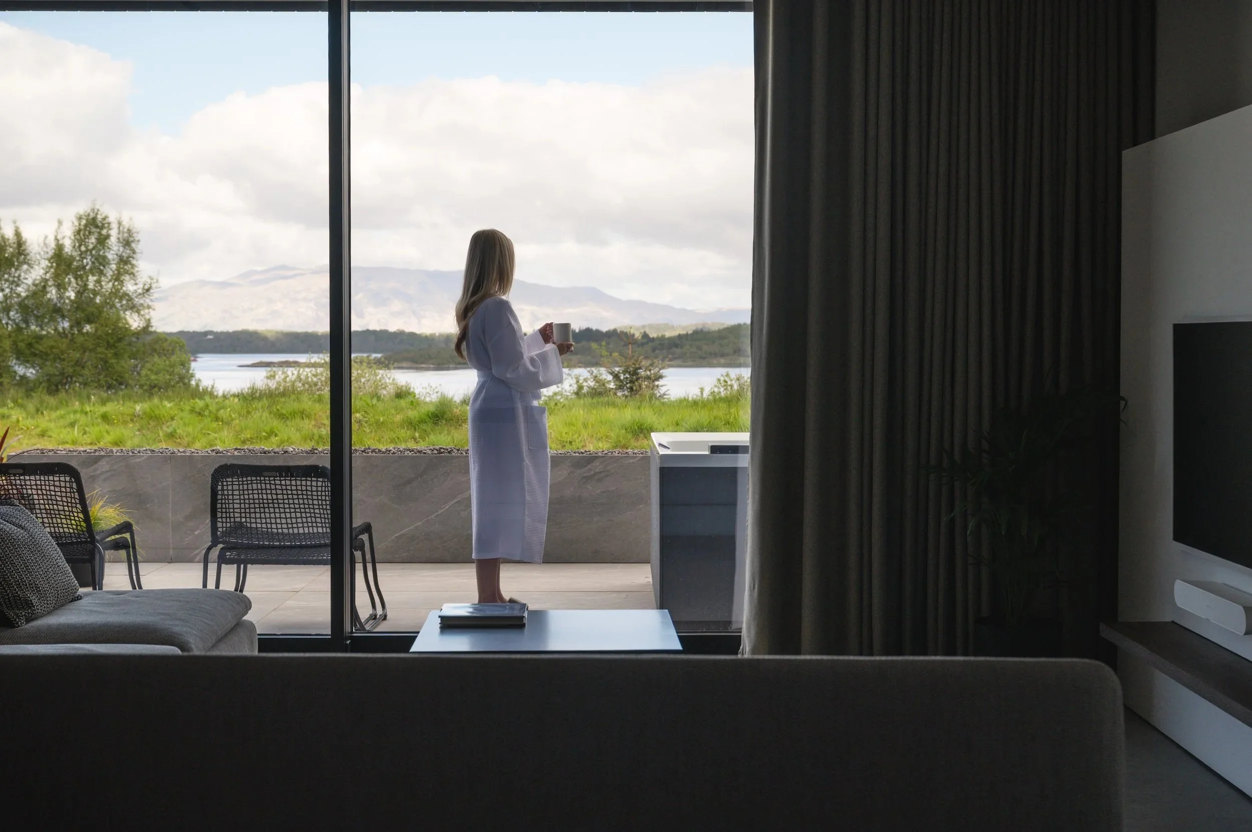A woman in a white bathrobe standing on a balcony with a coffee mug, overlooking a scenic landscape with trees, water, and mountains under a partly cloudy sky.