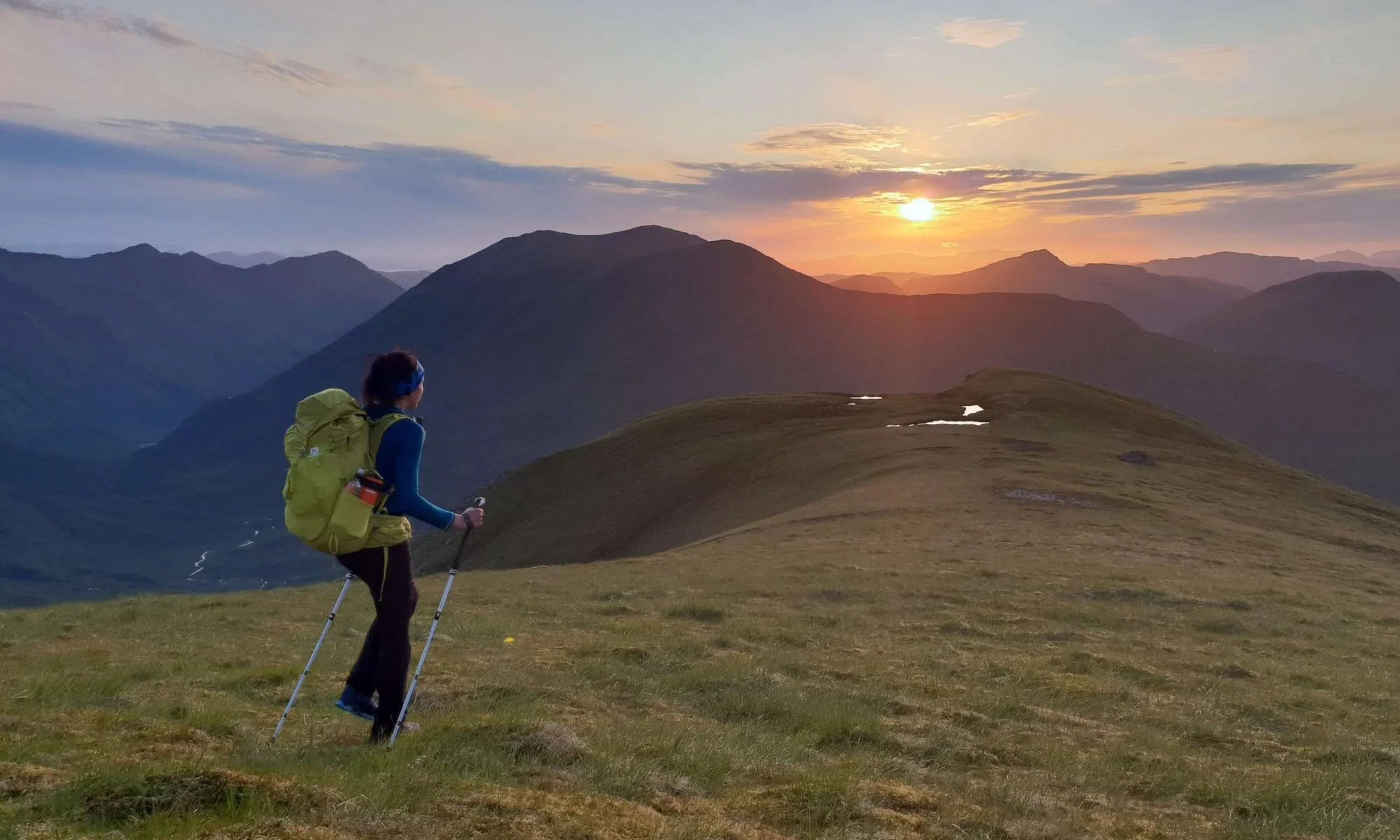 A person hiking with a yellow backpack and trekking poles on a grassy hillside at sunset, with mountains in the background.