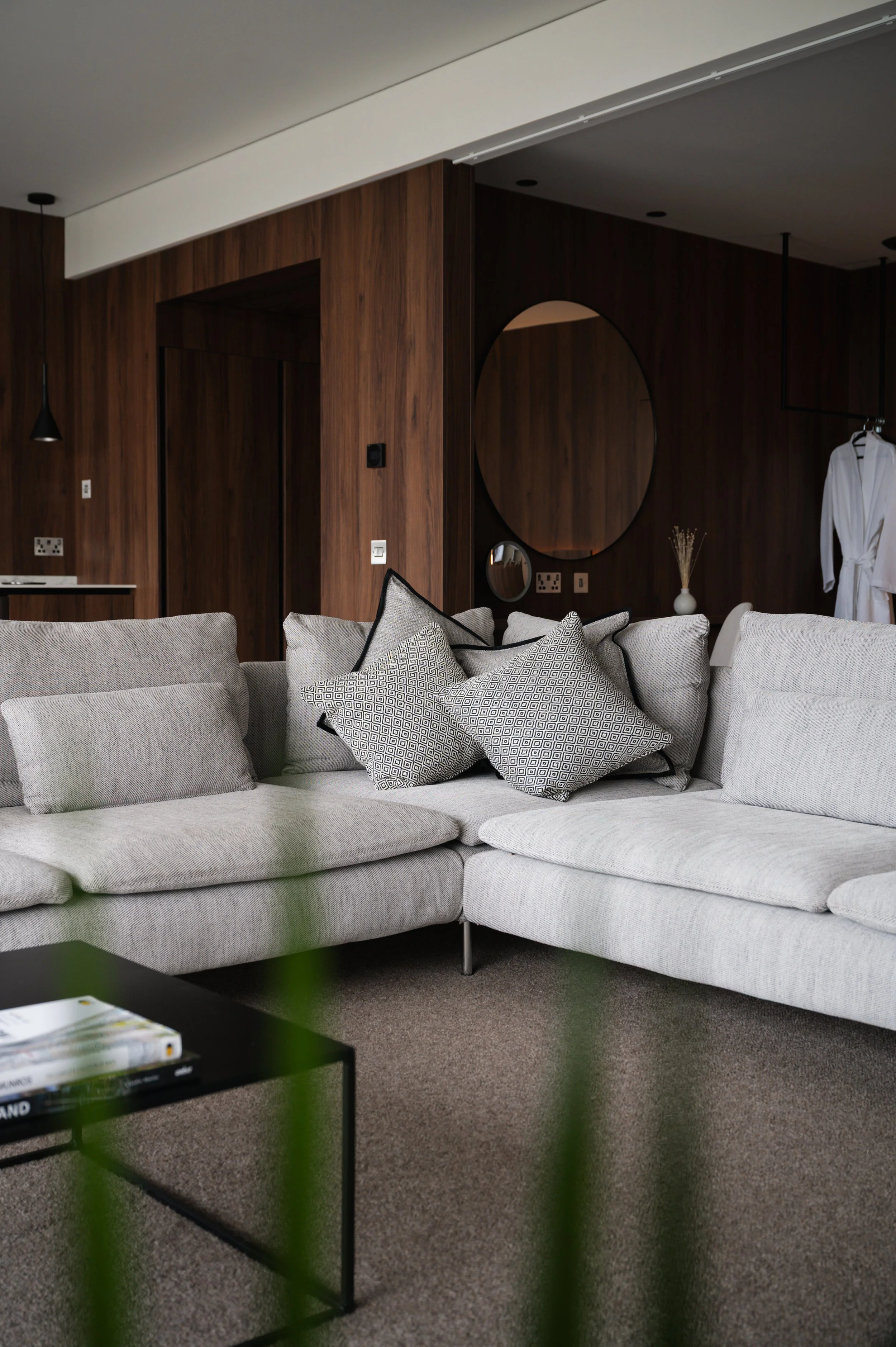 A modern living room with a curved light gray sofa decorated with black and white geometric-patterned pillows, a black metal coffee table with books, a large round mirror on a brown wood-paneled wall, white bathrobe hanging, a vase with dry flowers, and a small black pendant light.
