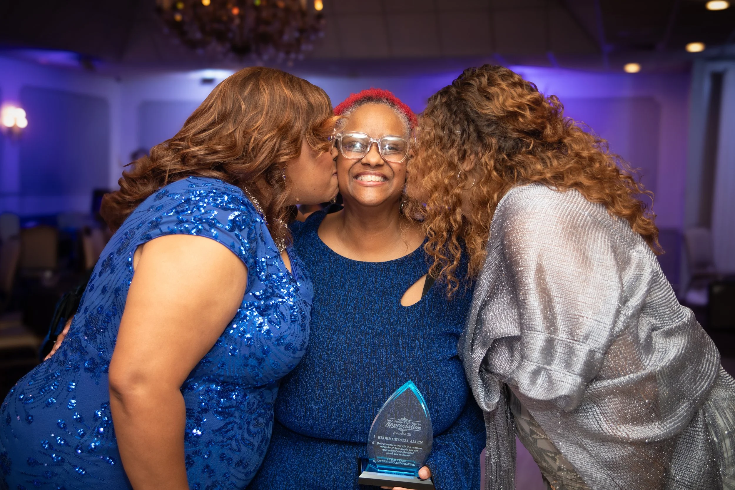 A woman in the center is holding an award and smiling as she is kissed on both cheeks by two women, one on each side, at an indoor event with purple lighting.