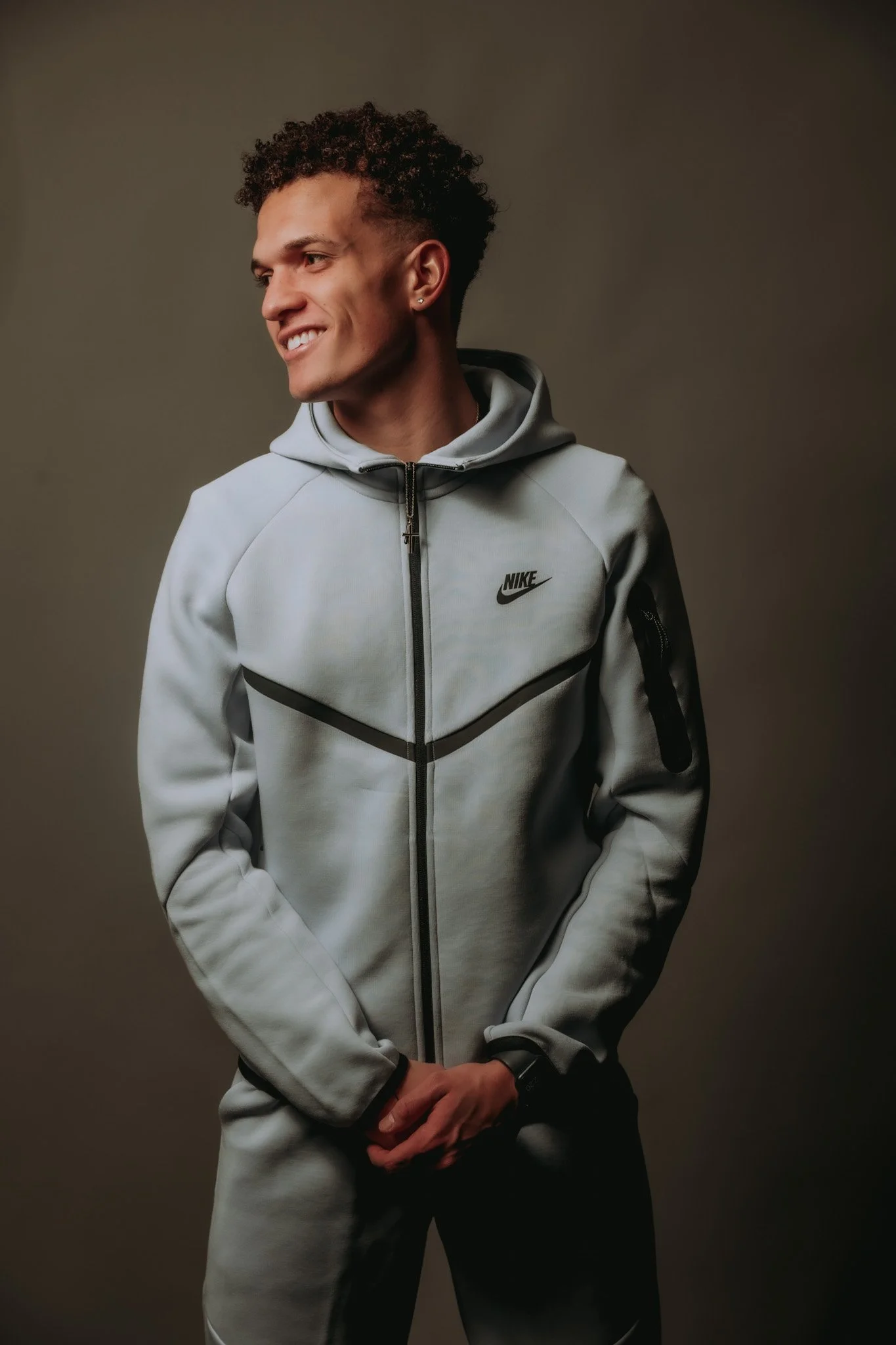 A young man with curly hair and earrings wearing a light gray Nike zip-up jacket and standing against a plain background.