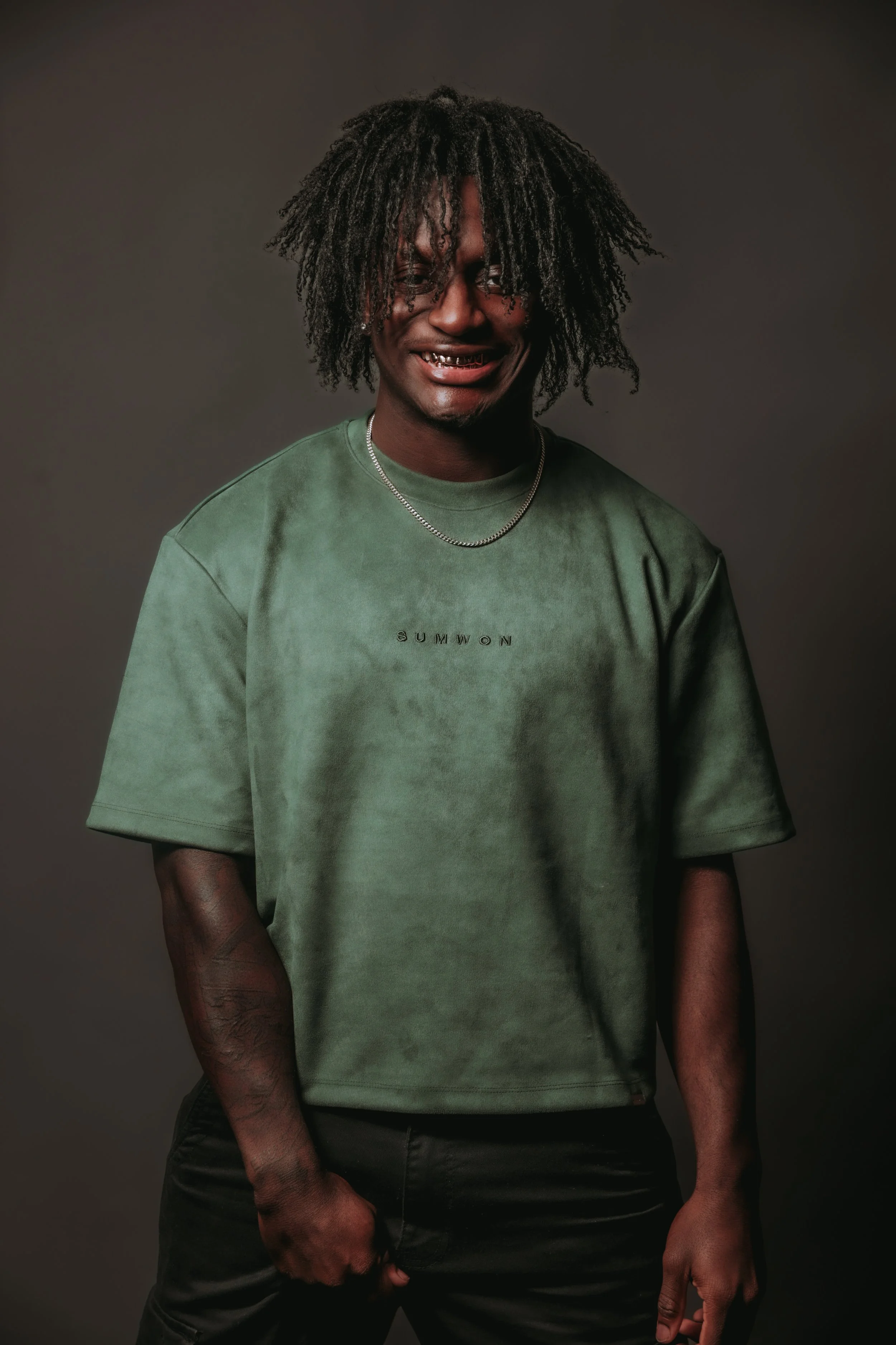A young man with dreadlocks, wearing a green T-shirt with the word 'SUMMON' and a silver chain, smiling with gold teeth, posing against a dark background.