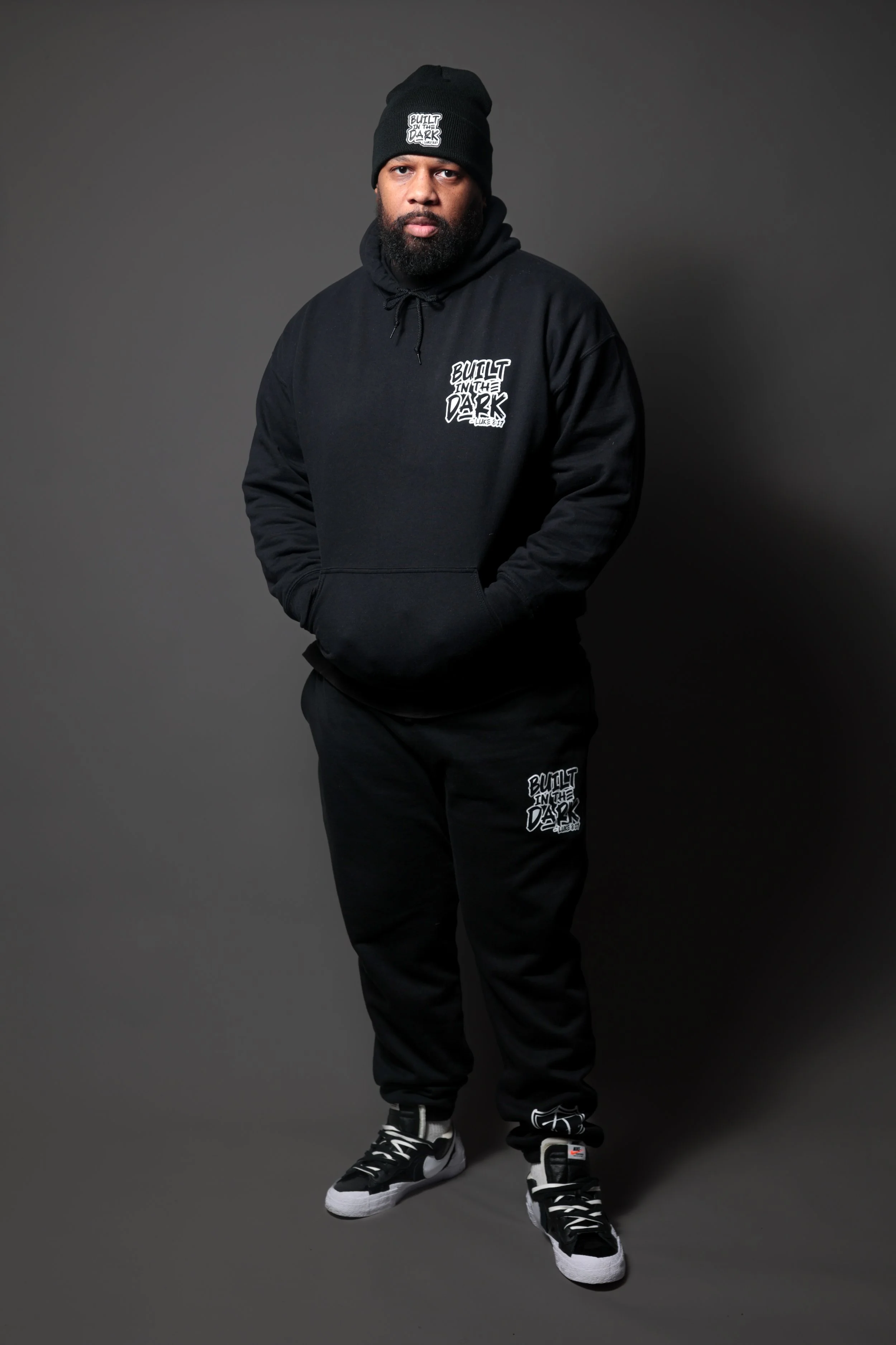 Man wearing a black hoodie, sweatpants, beanie, and sneakers, standing against a gray background. The hoodie and sweatpants have white text that says "Built in the Dark."
