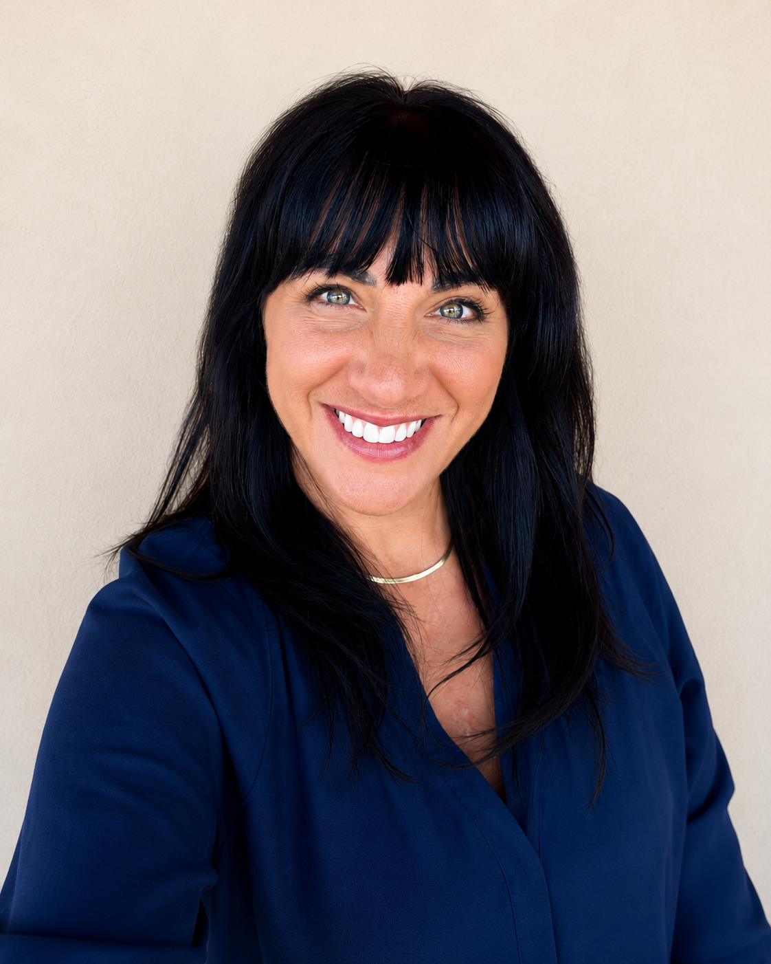 A woman with black hair and blue eyes smiling, wearing a navy blue top and a silver necklace, against a beige background.