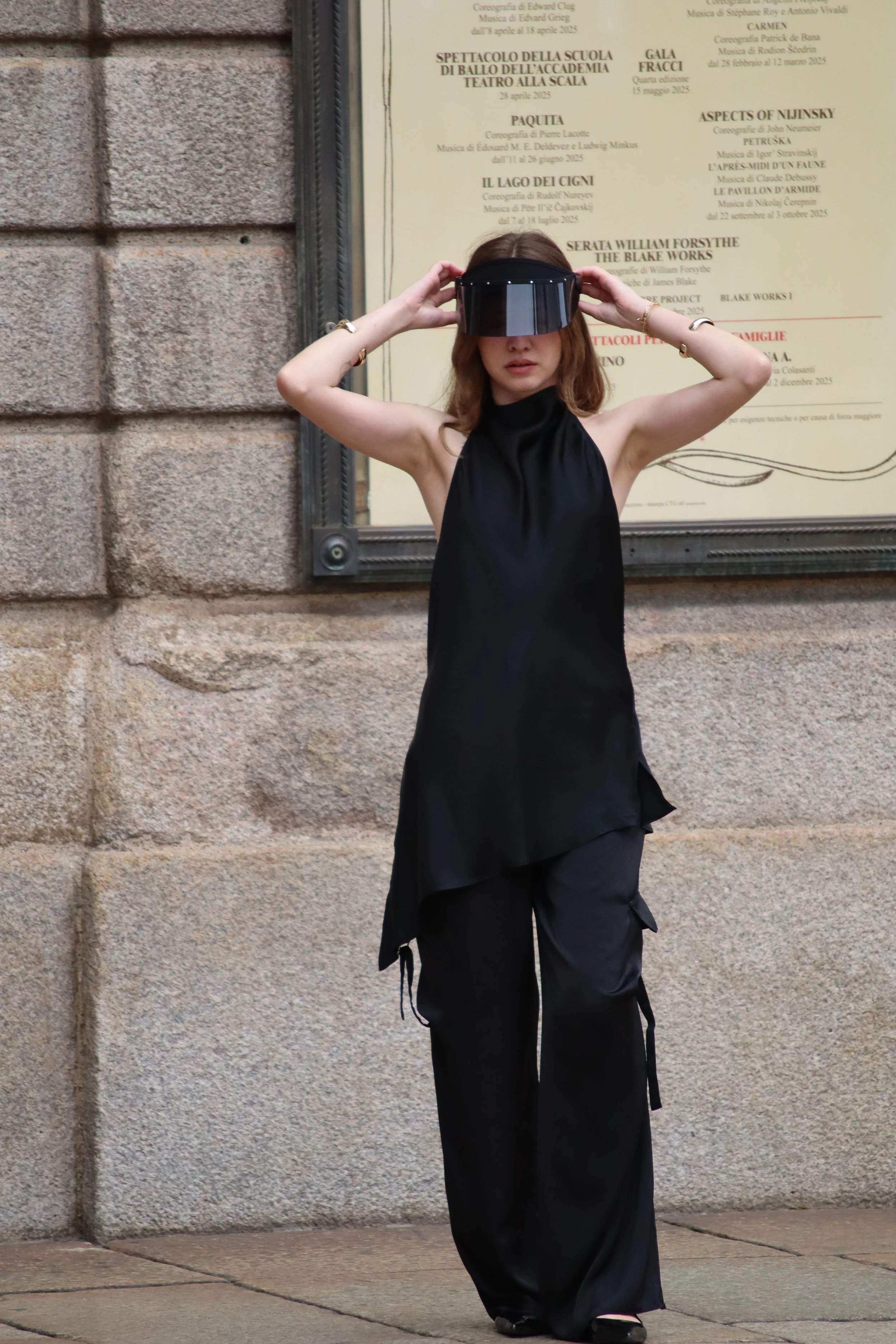 A woman in black clothing wearing a visor-style eyewear standing against a stone wall and a poster.