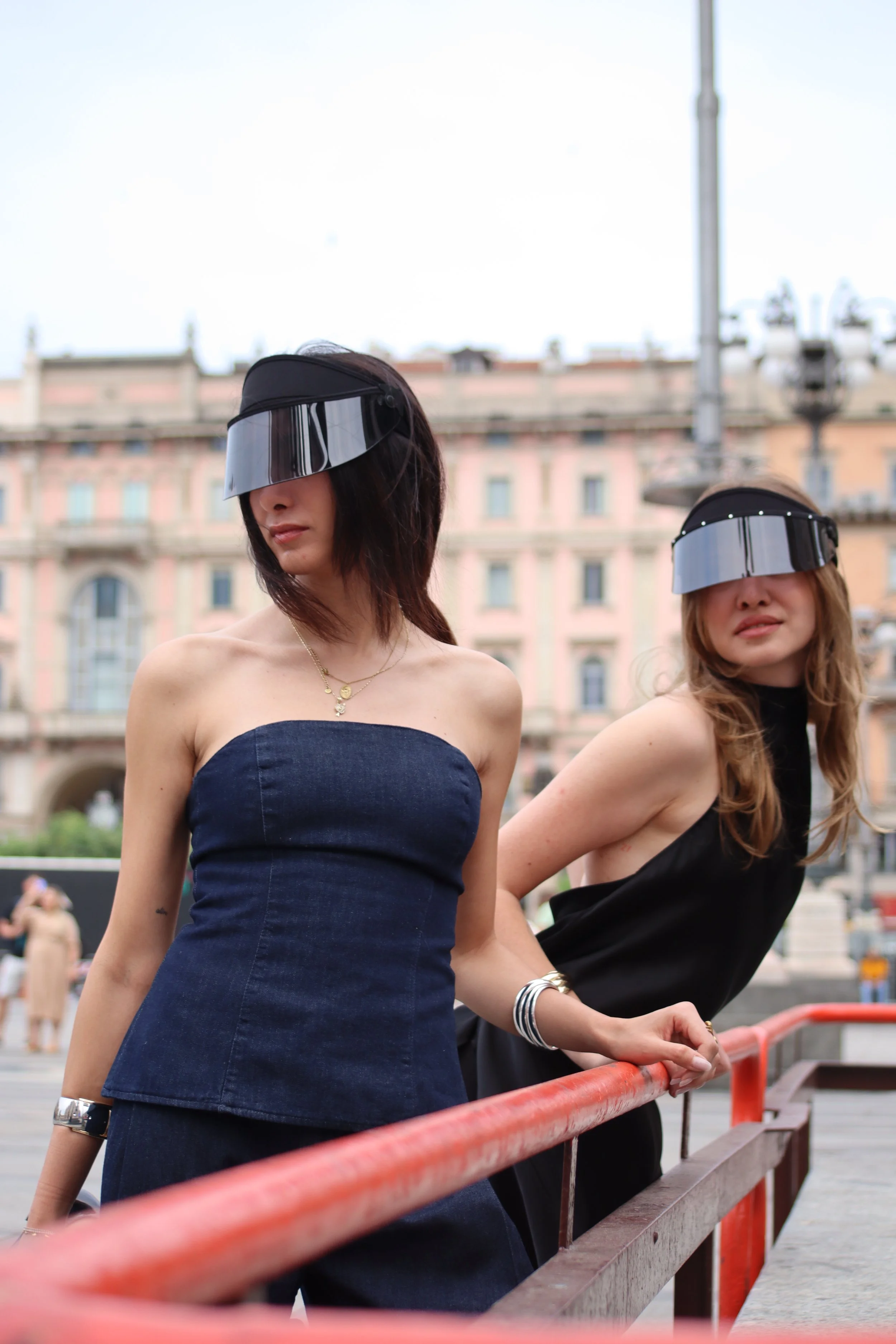 Two women wearing black visors stand outdoors in front of a historic building, dressed in stylish black and navy outfits.