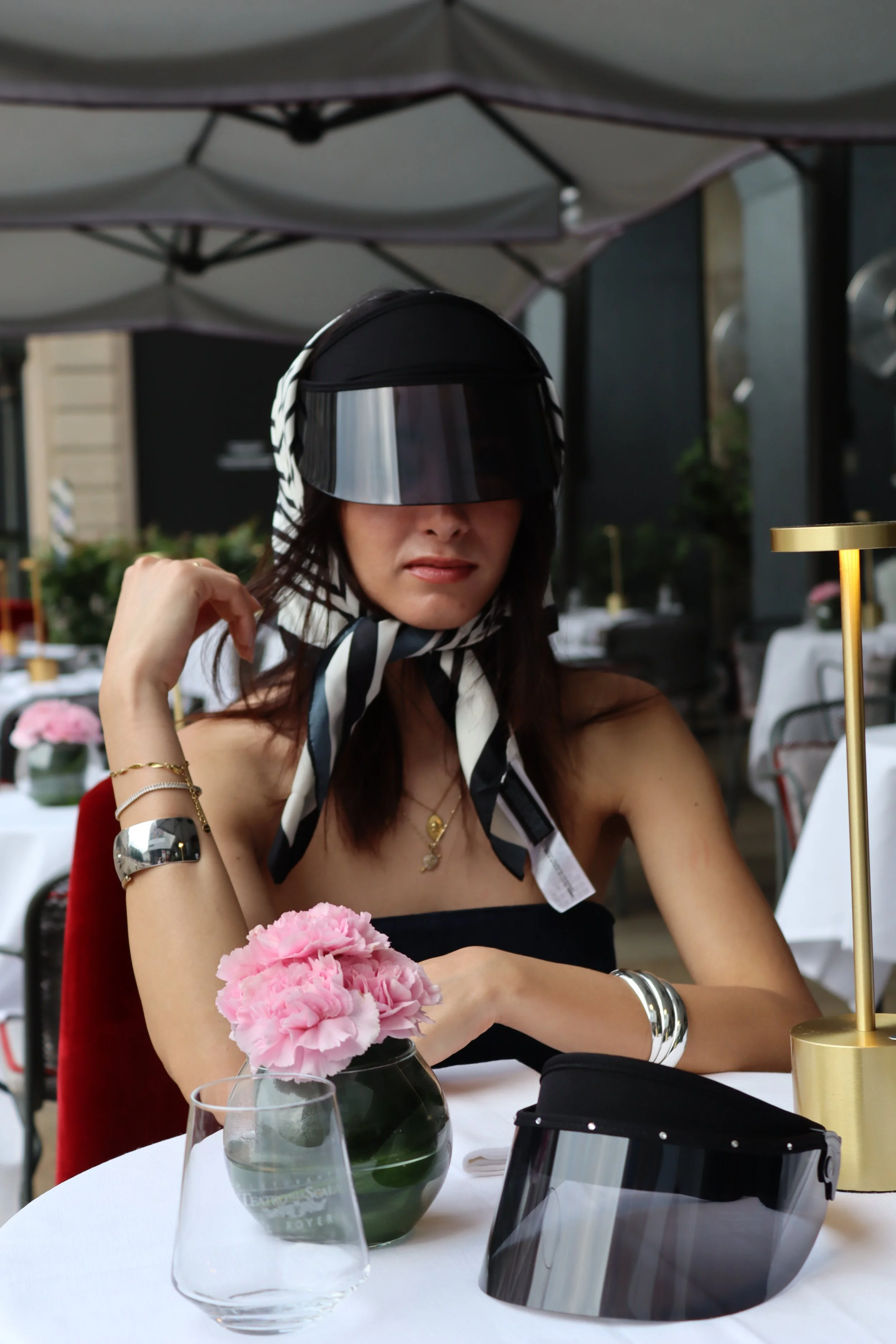 A woman wearing a black strapless top, large visor sunglasses, and a striped headscarf seated at a table in an outdoor restaurant. There are pink flowers in a glass vase, a black face shield, and a gold lamp on the table.