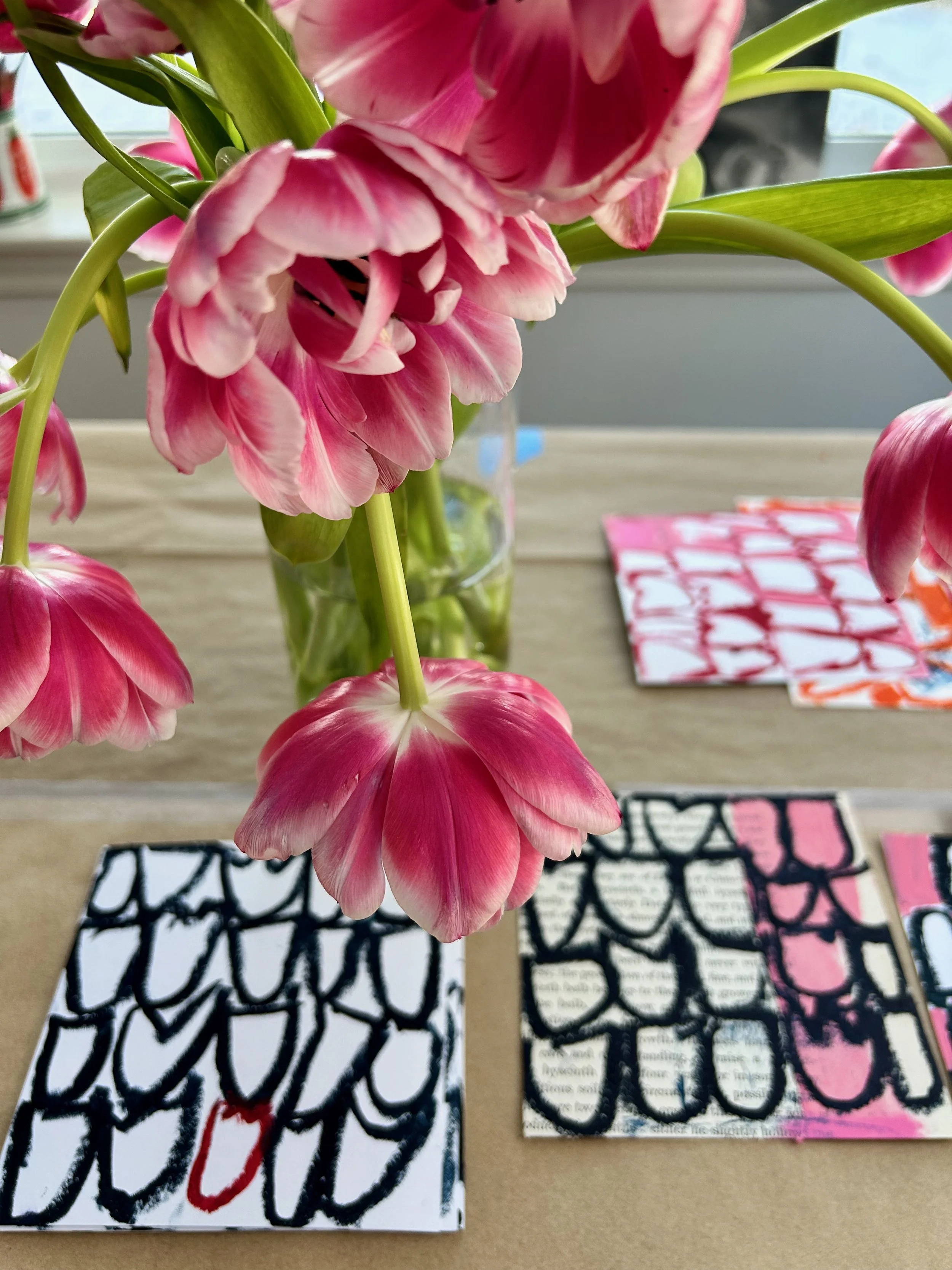 Close-up of pink and white flowers in a glass vase on a table, with colorful patterned papers with text underneath. Window sill in background.