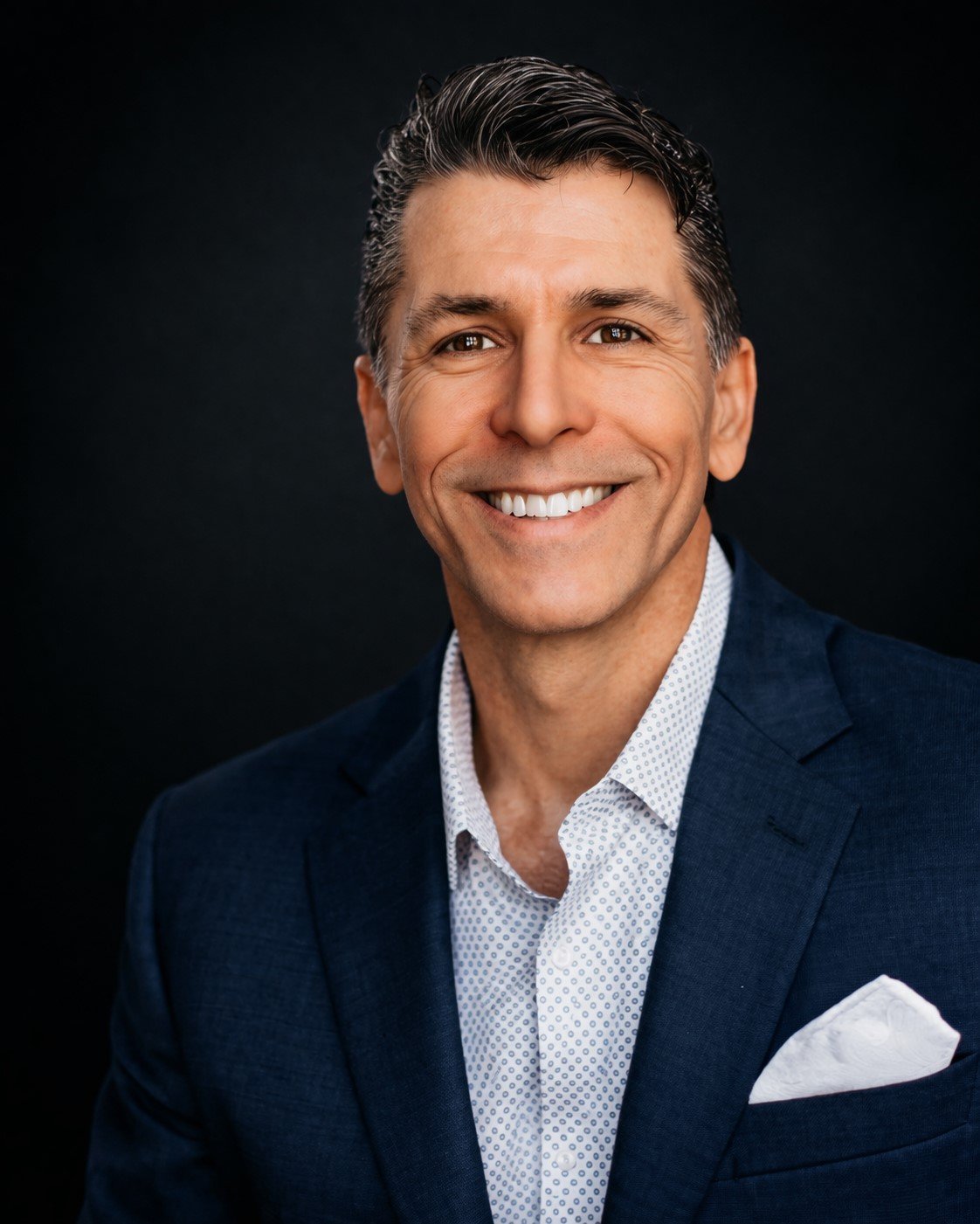 Close-up portrait of smiling middle-aged man with dark hair, dressed in a dark blue blazer, white patterned shirt, and white pocket square, against a black background.
