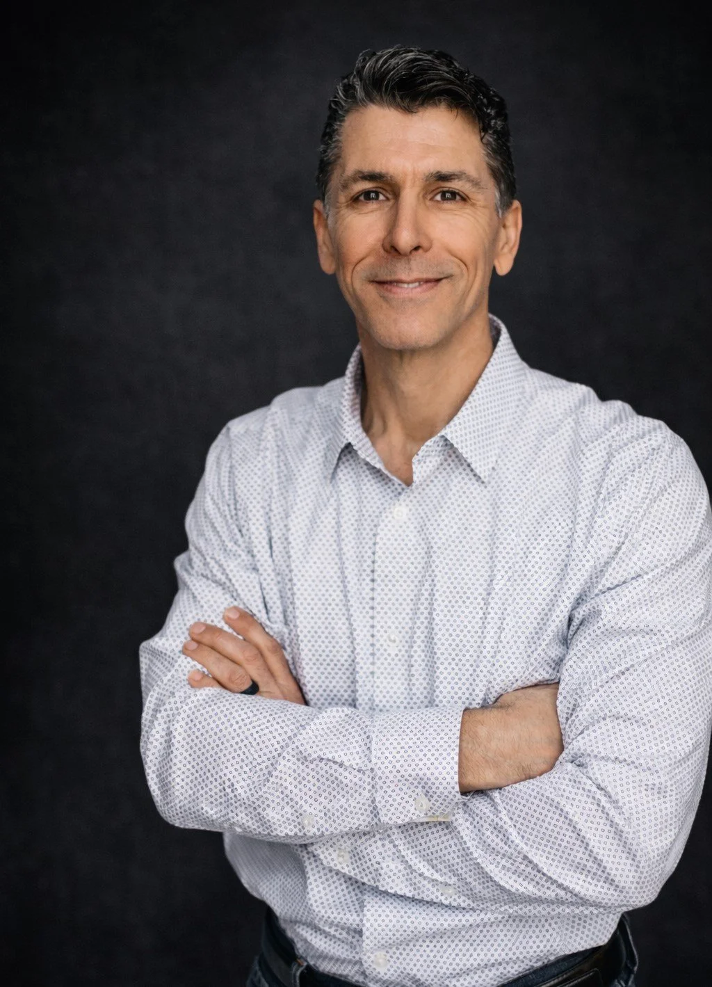 A smiling middle-aged man with dark hair, wearing a white patterned shirt, standing against a dark background with arms crossed.