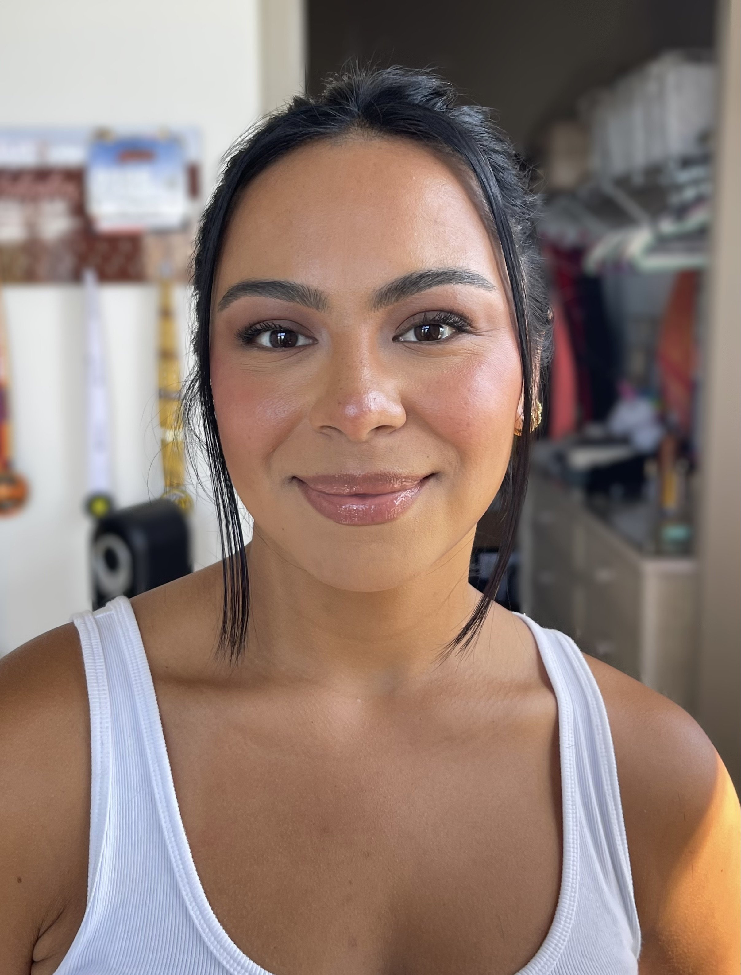 Close-up of a smiling woman with dark hair, wearing a white tank top, in a room with shelves and various items in the background.