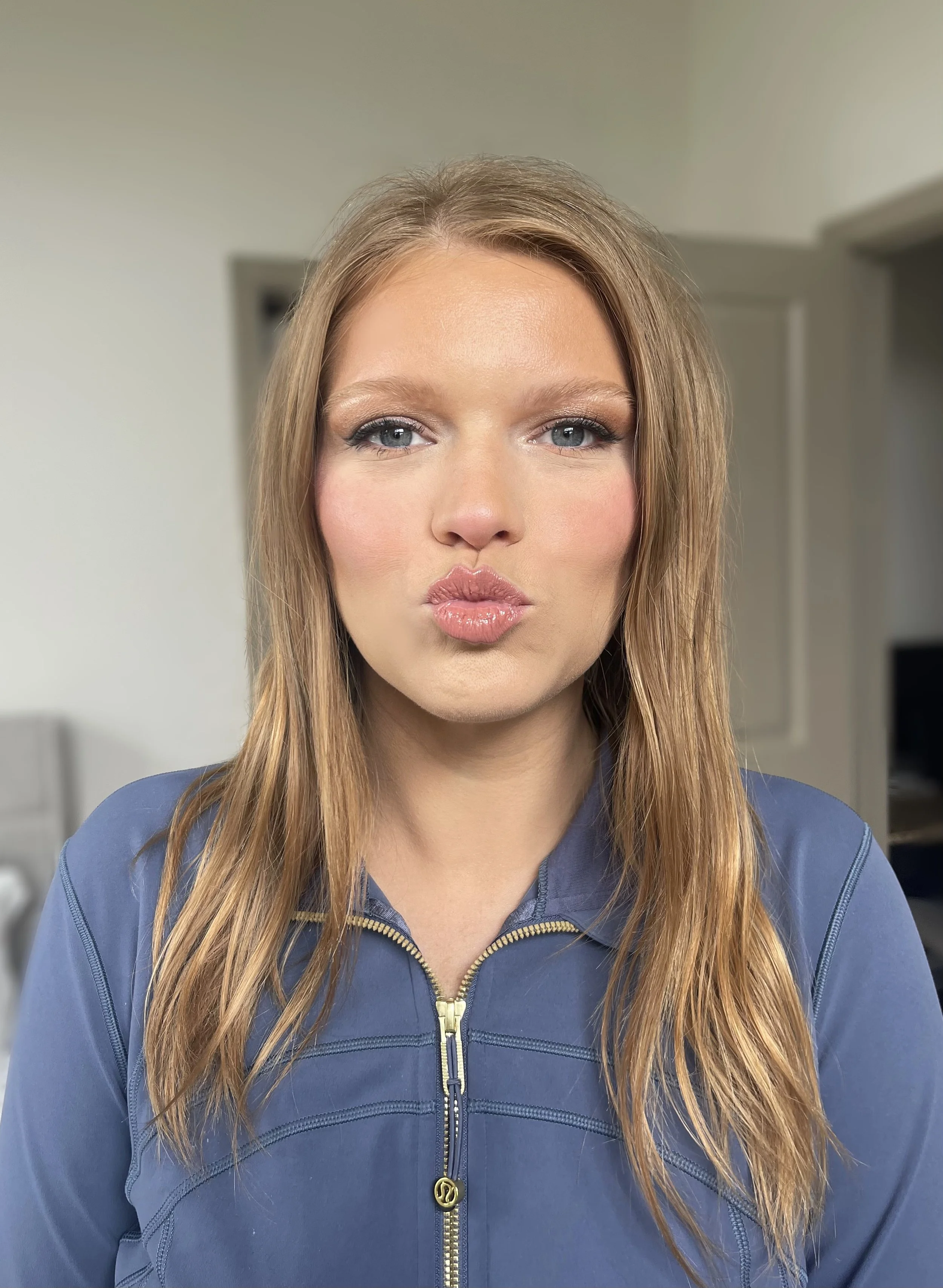 A woman with long, wavy blonde hair and blue eyes wearing a blue zip-up jacket, making a kiss face expression.