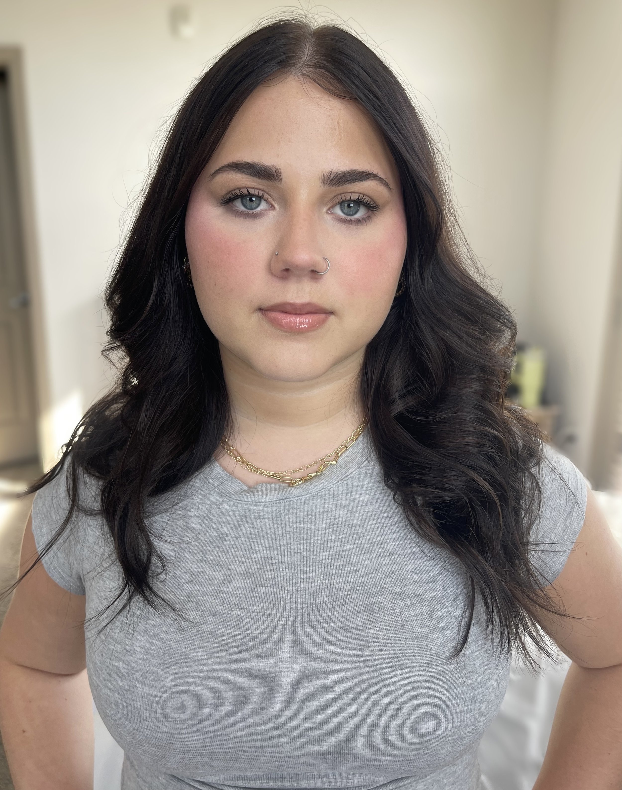 A woman with dark wavy hair, blue eyes, and light skin, wearing a gray t-shirt, gold necklaces, and small hoop earrings, standing indoors with a blurred background.