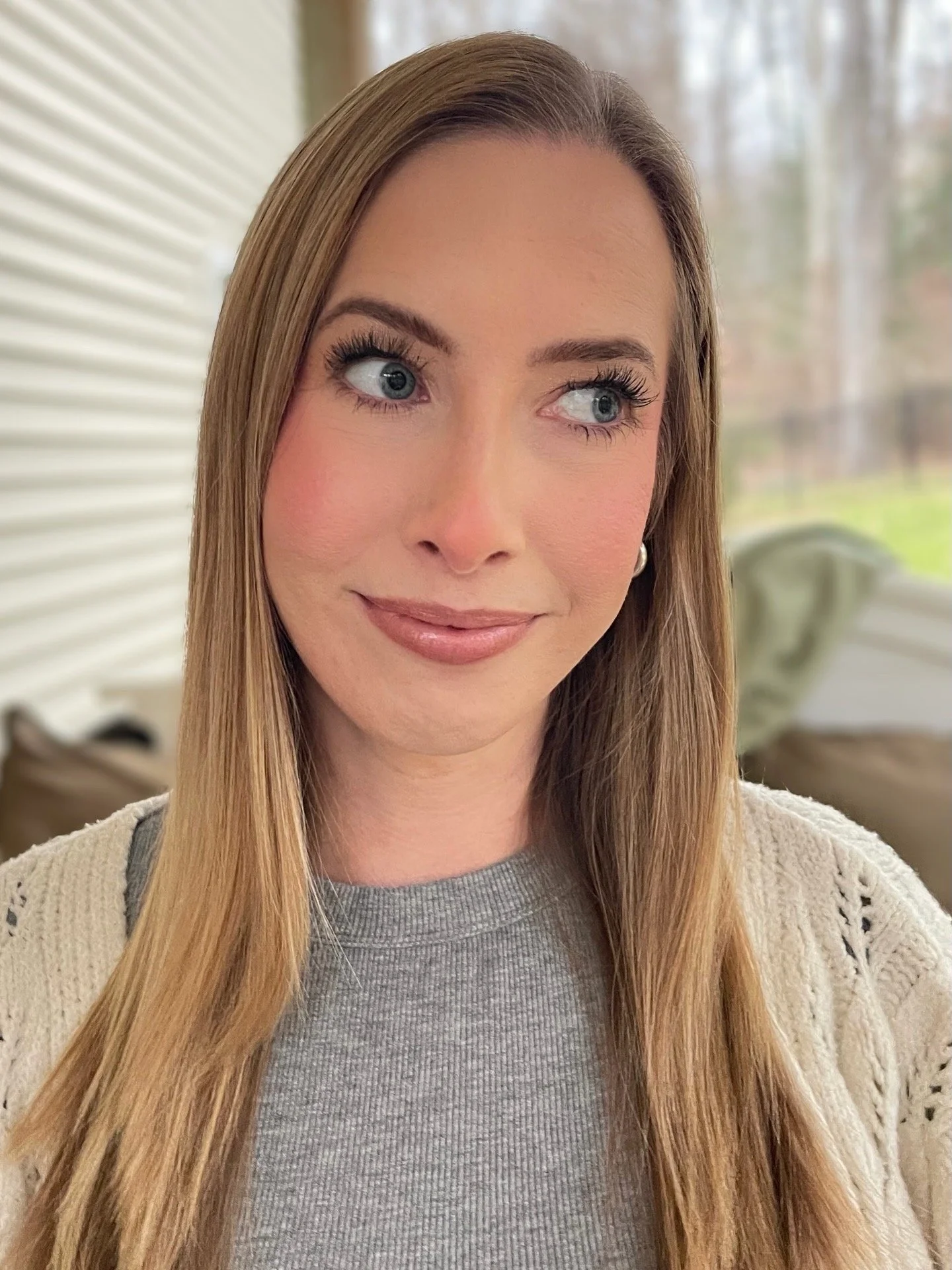A woman with long, straight blonde hair, blue eyes, and light makeup, wearing a gray top and cream knitted cardigan, standing indoors near a window with a blurred outdoor background.