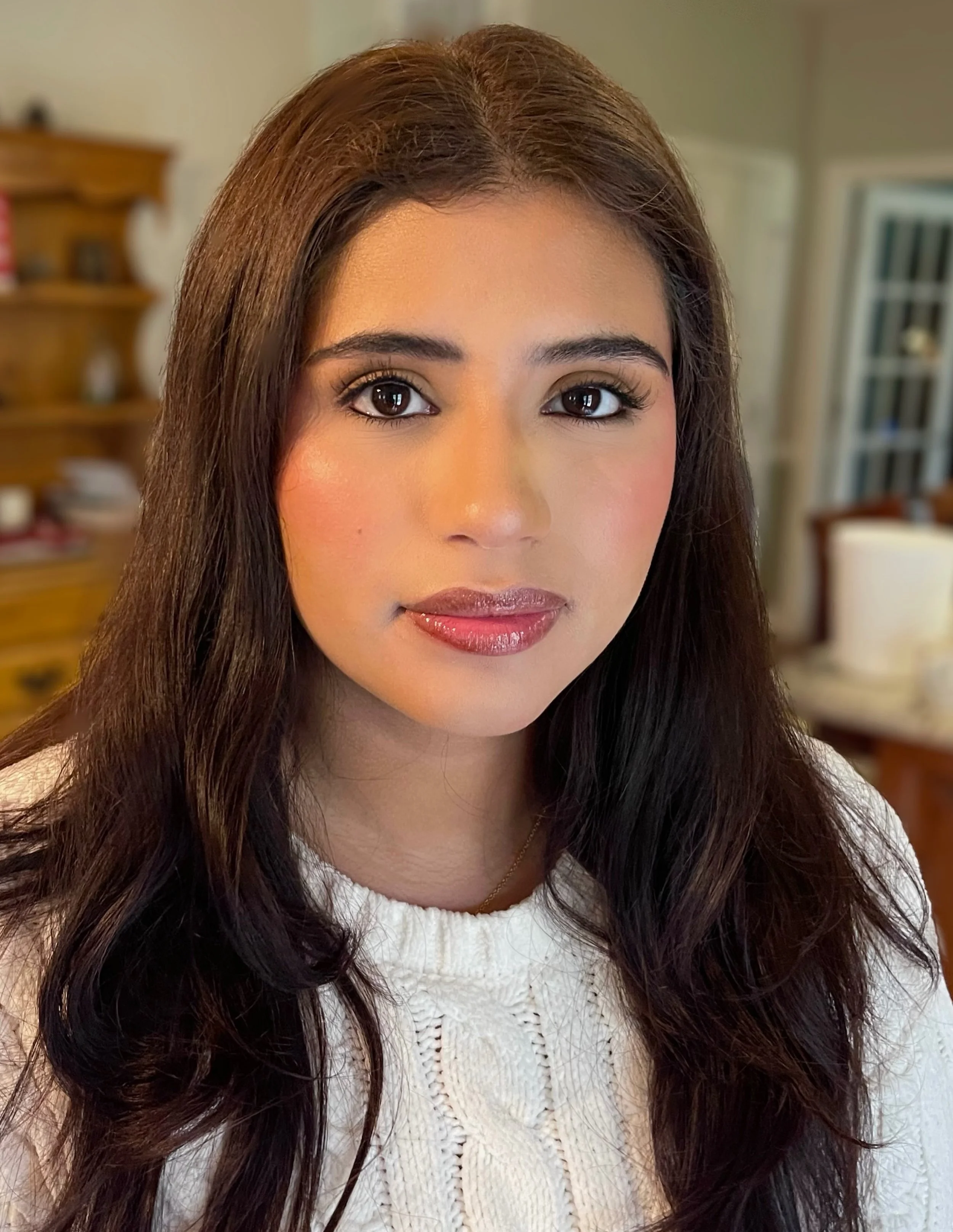 A woman with long, dark brown hair and makeup, wearing a white knitted sweater, standing in a kitchen or dining area.