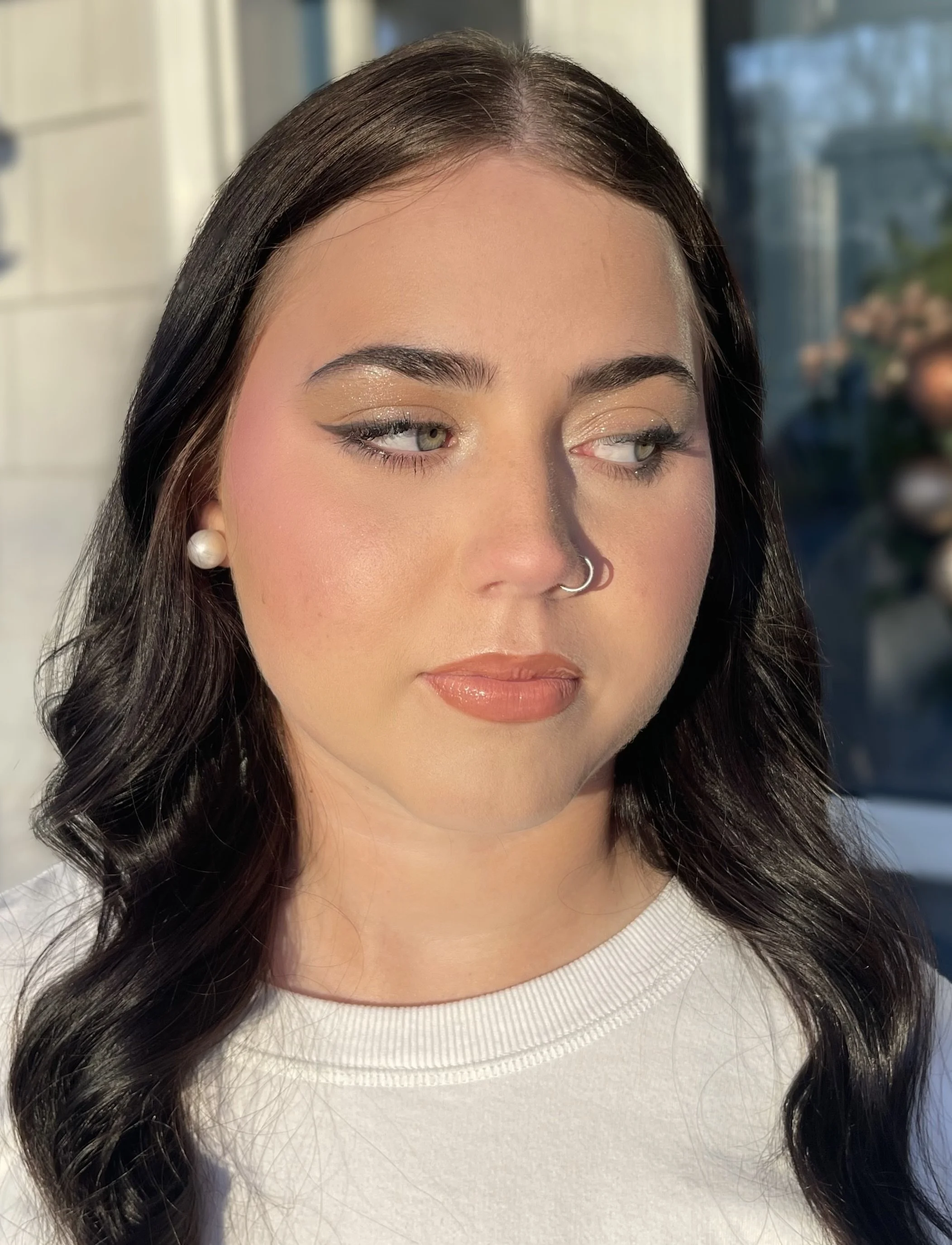 Close-up of a woman with dark hair, wearing pearl earrings and a nose ring, with makeup, looking slightly to the side, bathed in sunlight.
