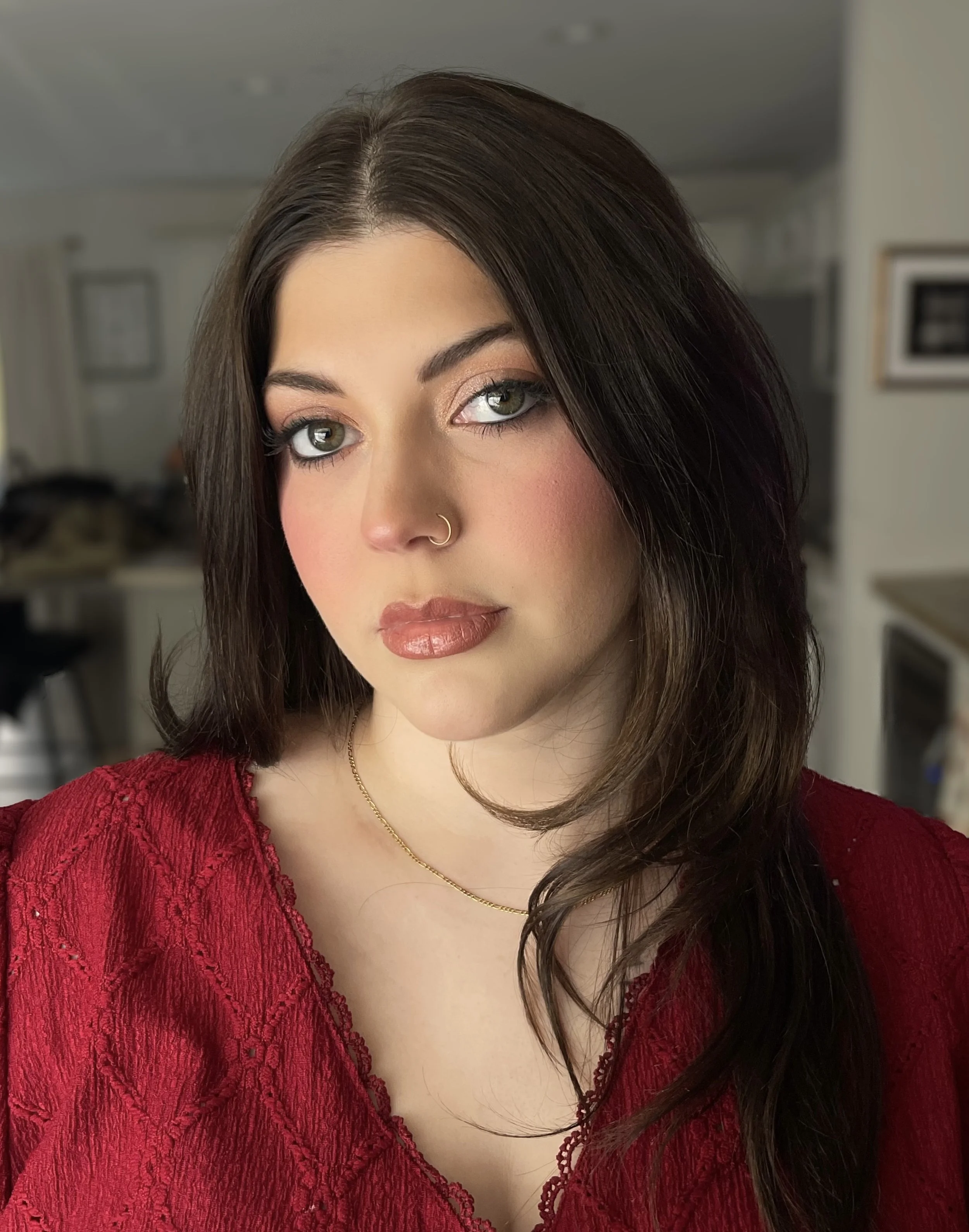 A woman with long dark brown hair and green eyes, wearing a red lace top and gold necklace, posed indoors with a blurred kitchen background.
