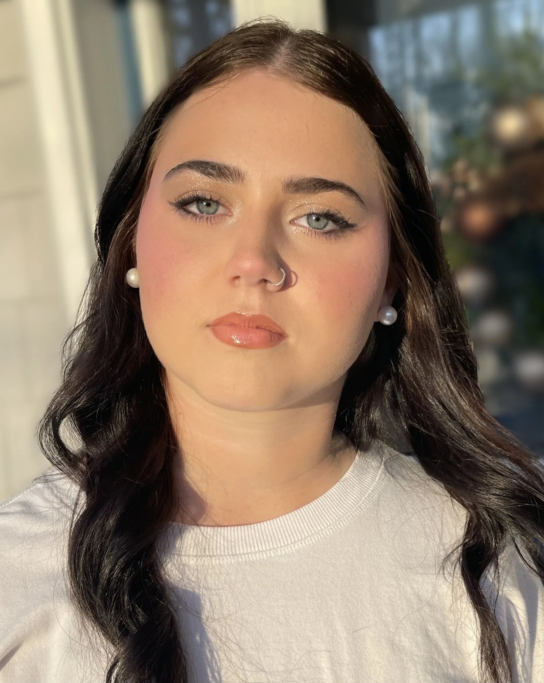 A woman with light skin, dark wavy hair, blue eyes, pearl earrings, wearing makeup with glossy lips, and a septum piercing. She is outdoors with sunlight on her face, standing in front of a blurred background with trees and buildings.