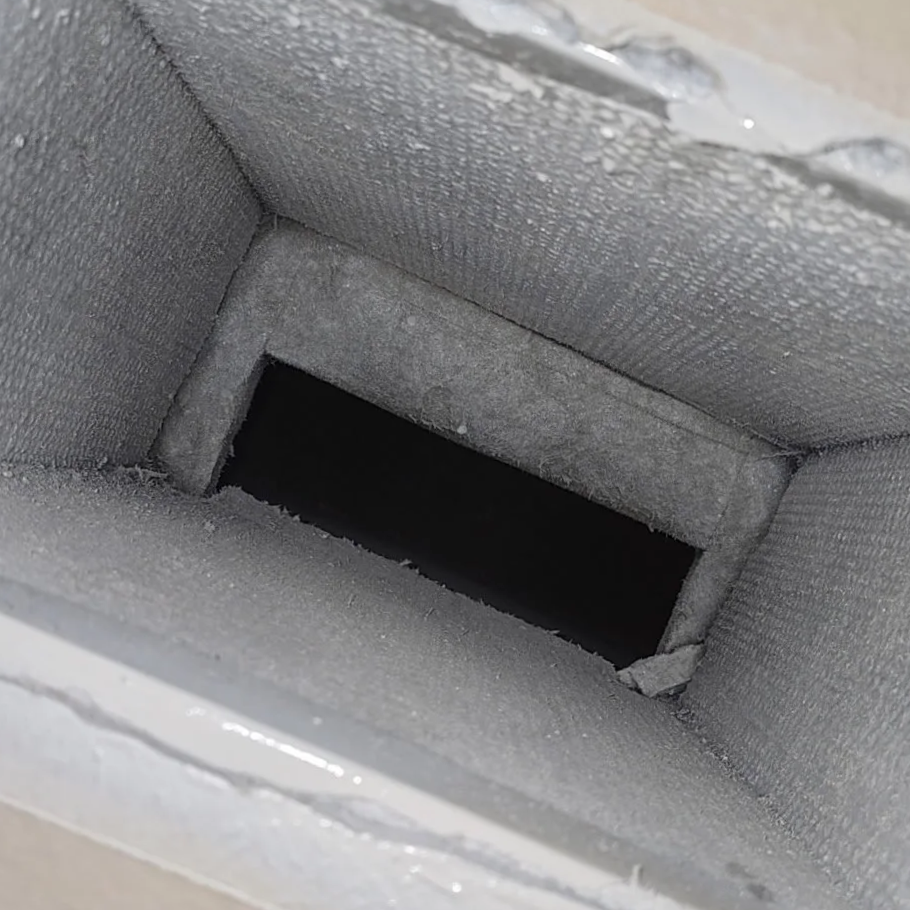 A close-up of a small, rectangular, dusty ventilation duct or vent with a dark opening inside.