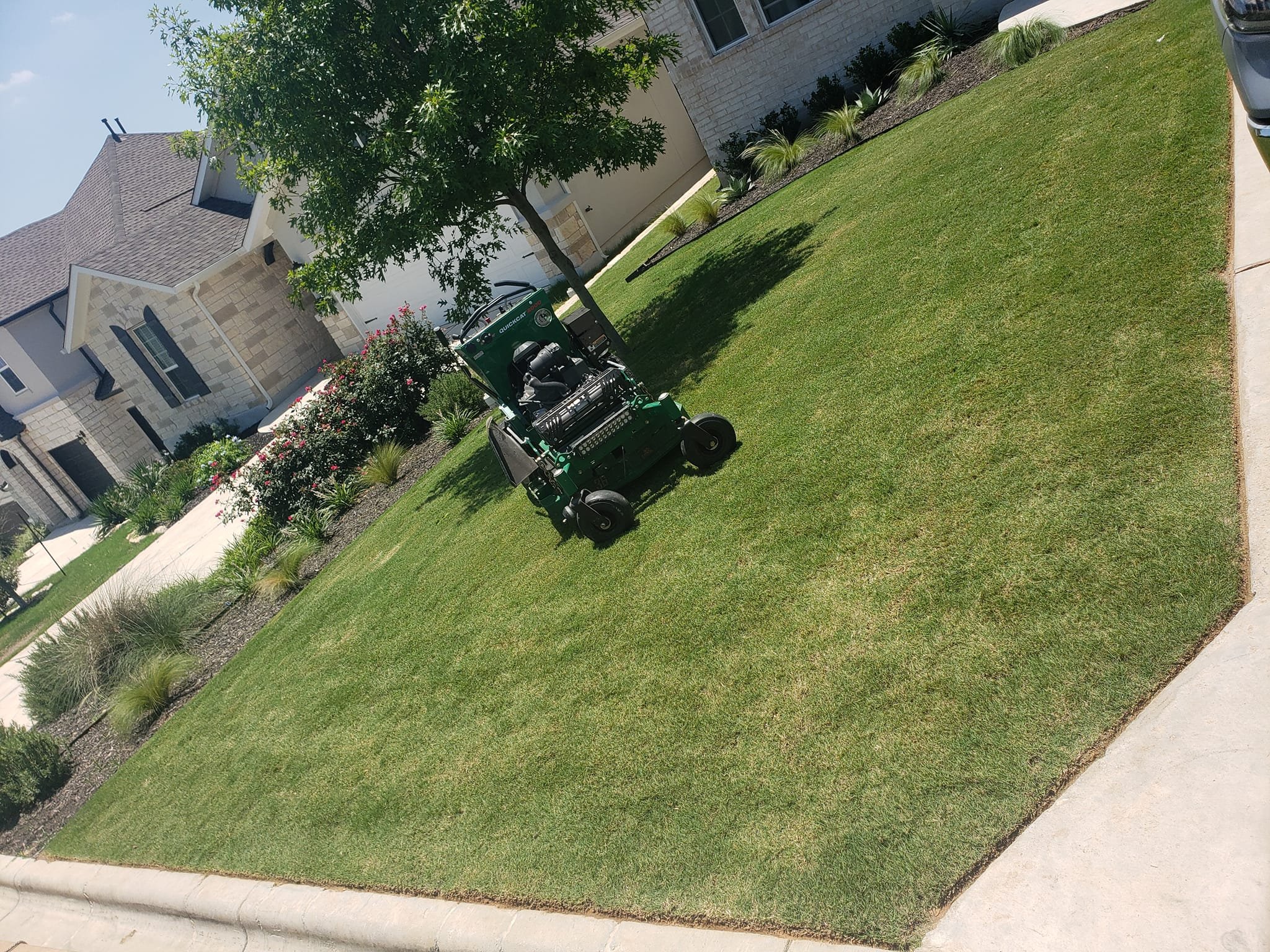 A small green robotic lawn mower on a well-maintained front yard next to a sidewalk, with a tree casting a shadow on the grass, in front of a modern house with stone and white siding.