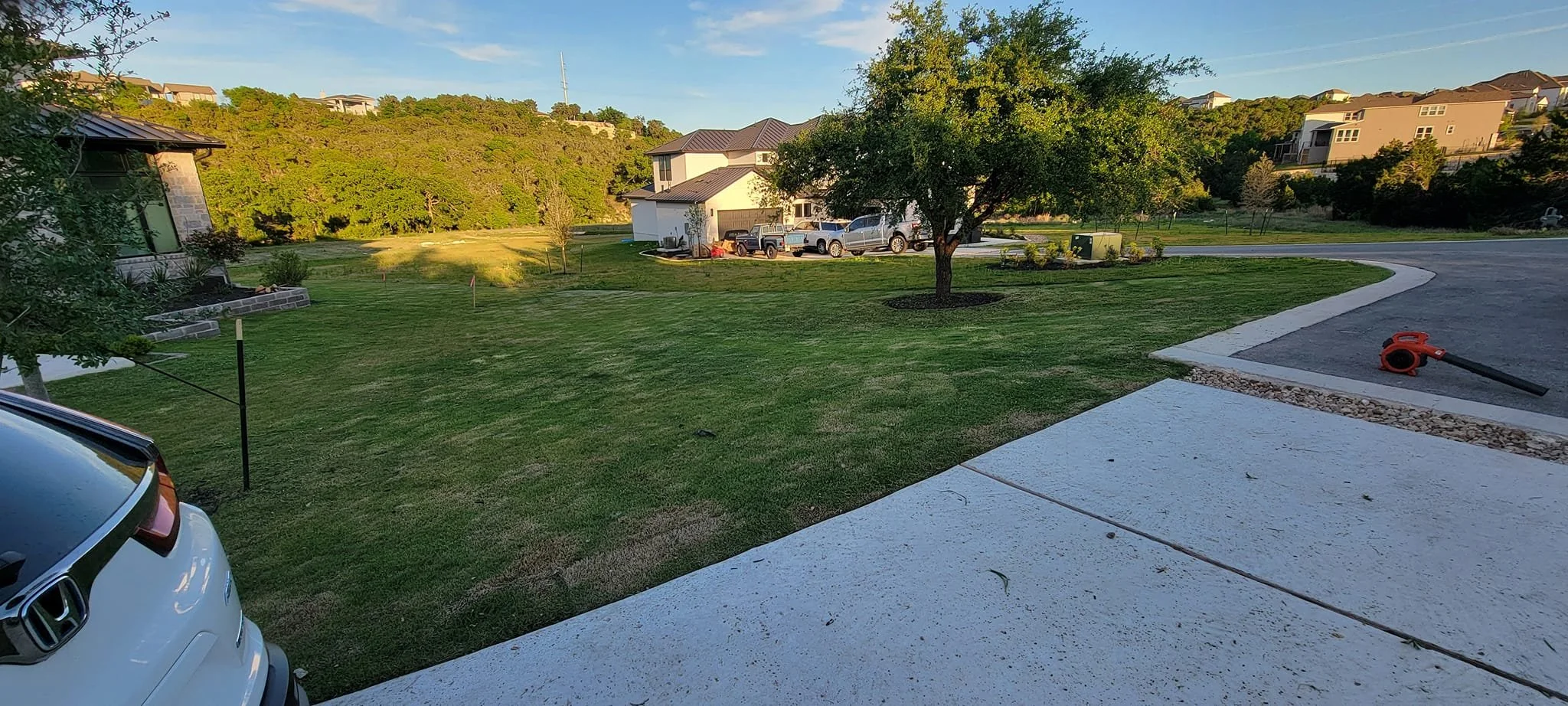 A suburban neighborhood with green lawns, trees, and houses in the distance. A concrete sidewalk and driveway are visible, with a leaf blower on the driveway. Several parked trucks are near a white house, with a mountain or hill covered in trees in t