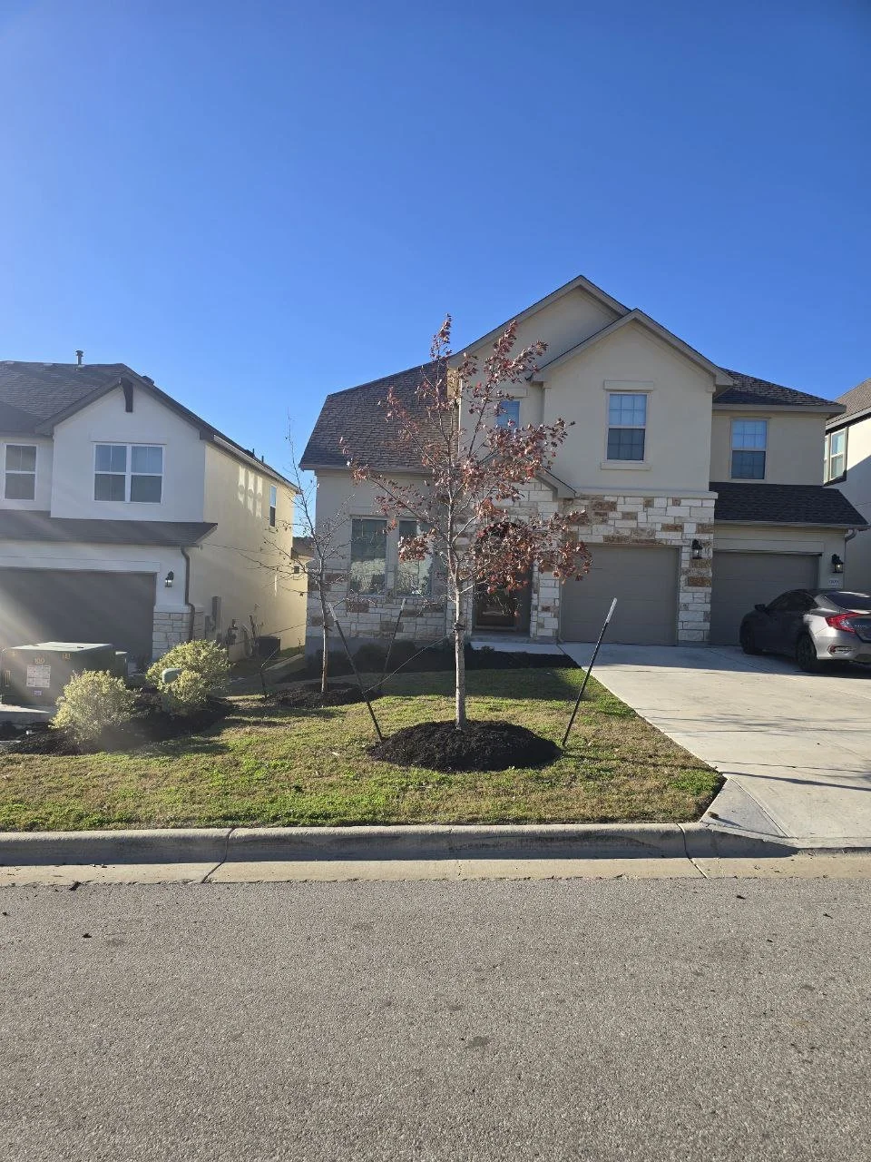 A two-story house with a yellow exterior, stone accents, and a dark roof. In front, there is a small lawn with a newly planted tree supported by stakes. The driveway has a car parked on the right, and a sidewalk with a curb lines the street.