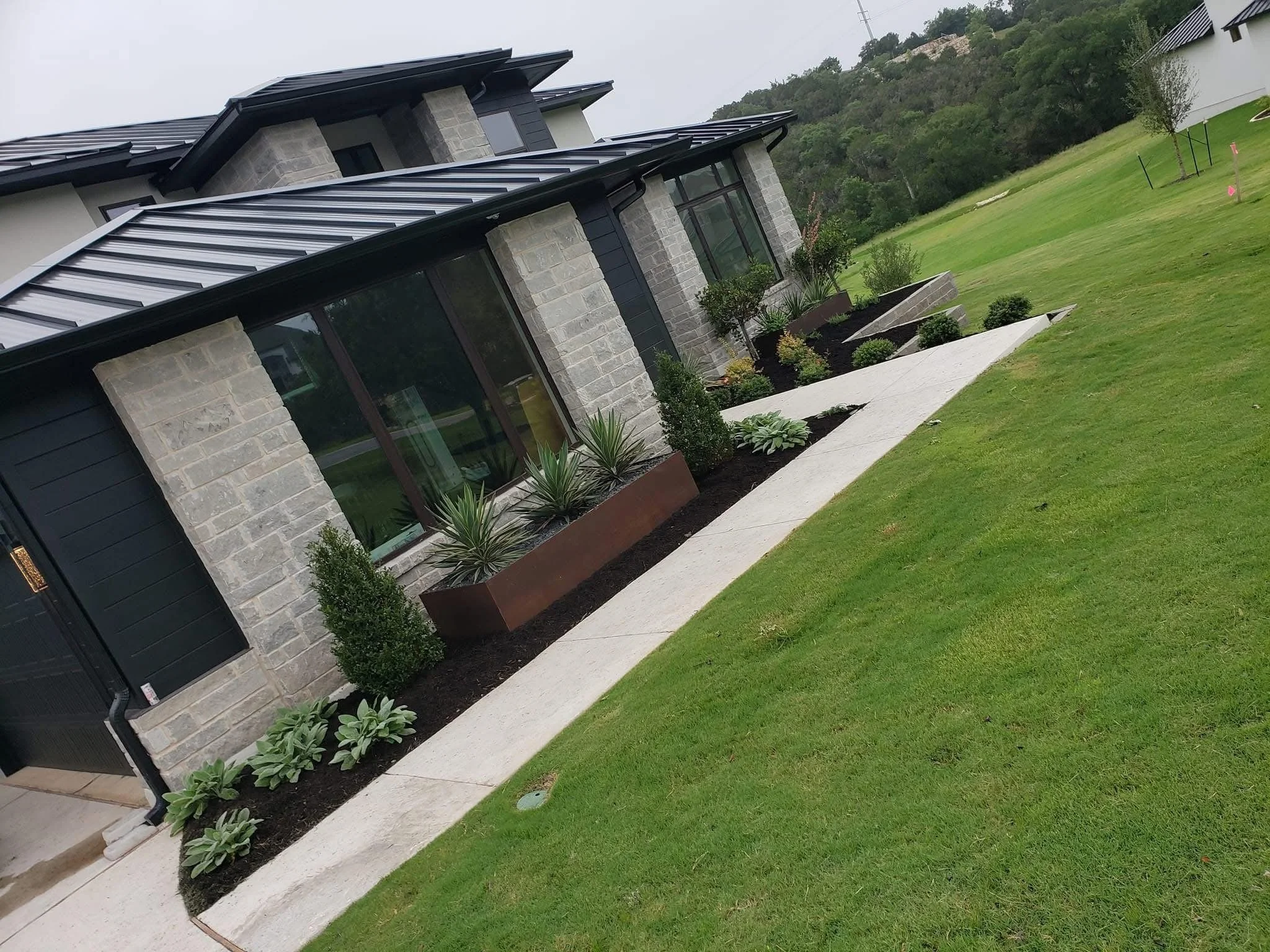 Modern residential house with stone and black siding, large windows, and metal roof, surrounded by landscaped garden beds with plants and shrubs, and a concrete walkway on a grassy lawn