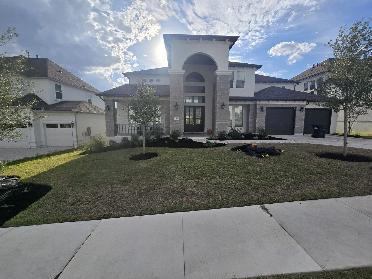 Front view of a modern two-story house with a large arched entryway, stone accents, and three garage doors, with a front lawn, trees, and a partly cloudy sky.