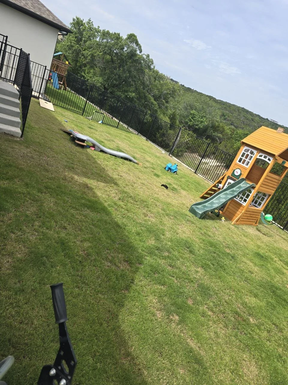 A backyard with a green playhouse with a slide, a small blue toy car, and a playground set with a slide and climbing area. The yard is enclosed with a black fence, with a house on the left and trees in the background.