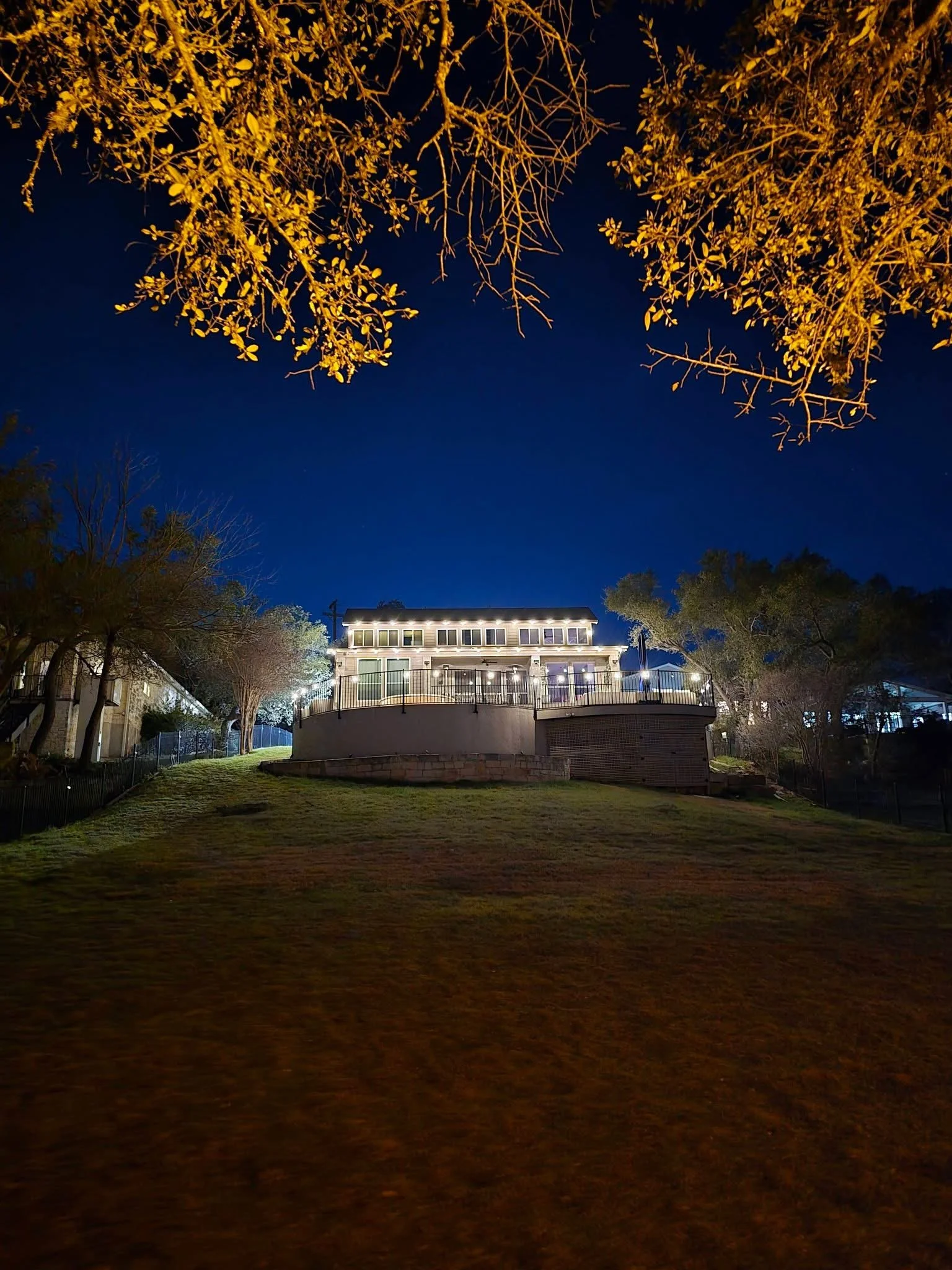 A house with lights on at night, located on a hill with a well-kept lawn, trees on either side, and a dark blue sky overhead.