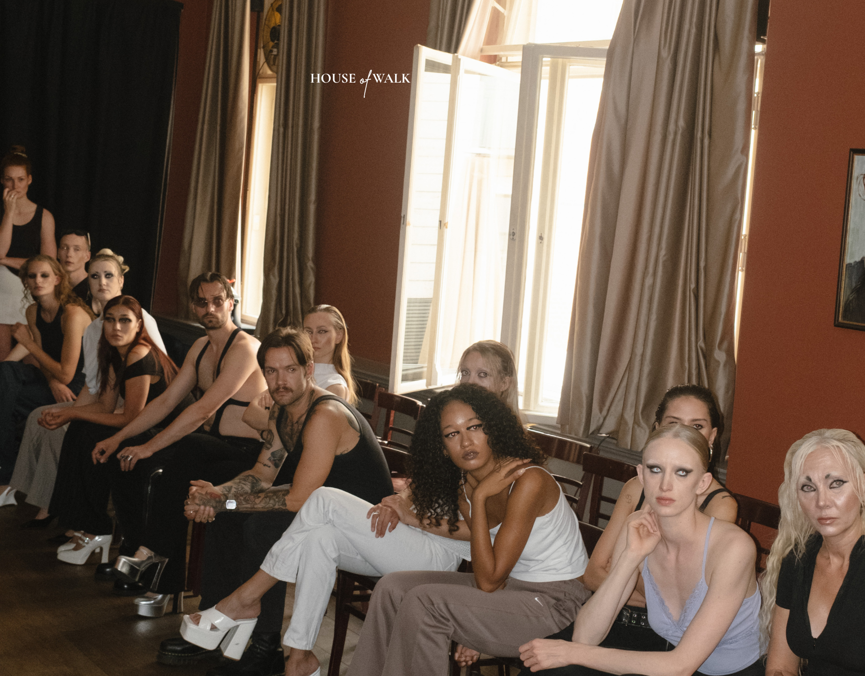 Gruppe von Menschen, die in einem Raum sitzen, einige mit Make-up und unterschiedlichen Stilen, vor einem offenen Fenster mit weißen Vorhängen, im Hintergrund schwarze Vorhänge und ein Gemälde an der Wand.