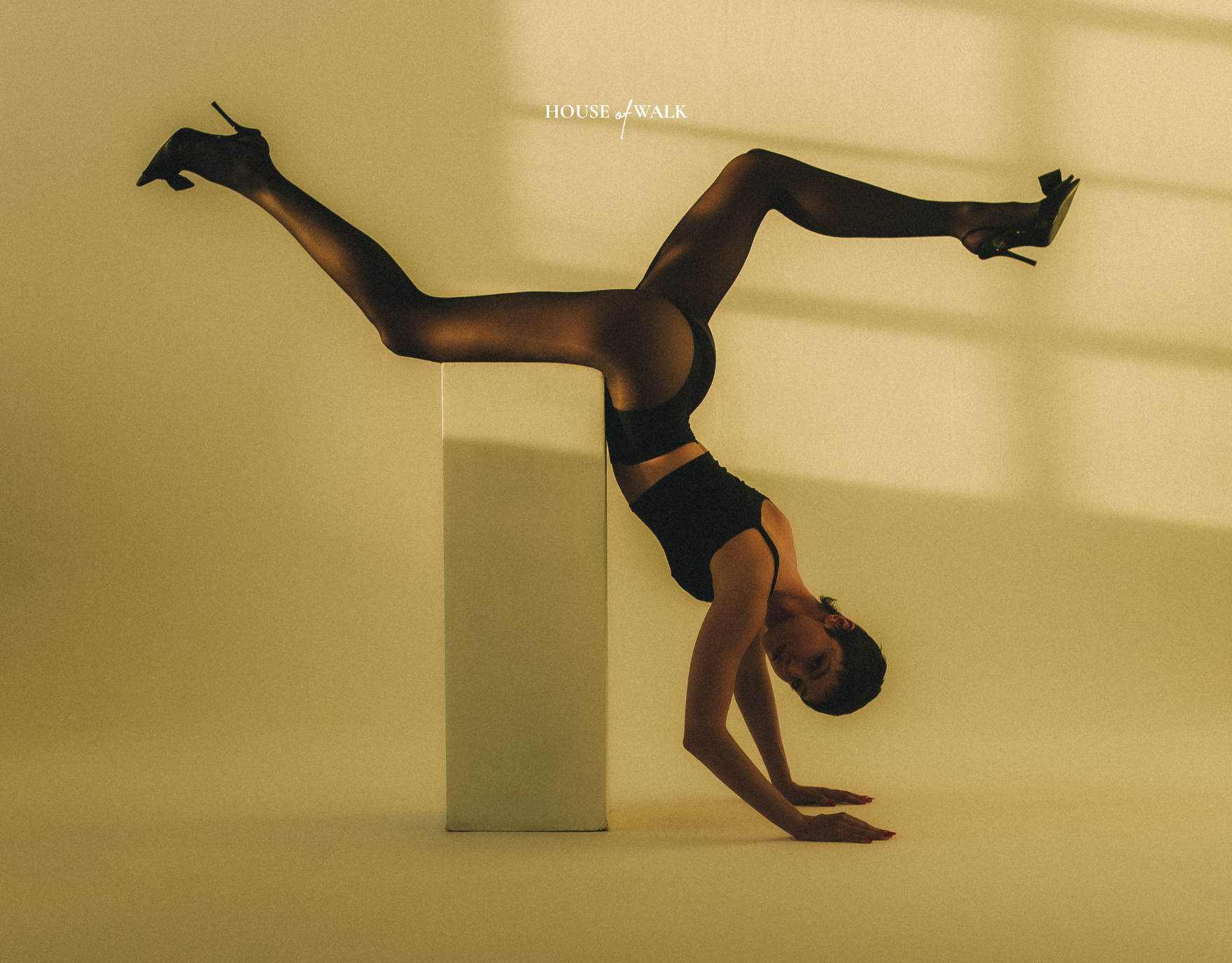 Frau in schwarzen Strümpfen und High Heels macht einen Handstand, mit den Beinen auf einem erhöhten Block, auf gelbem Hintergrund