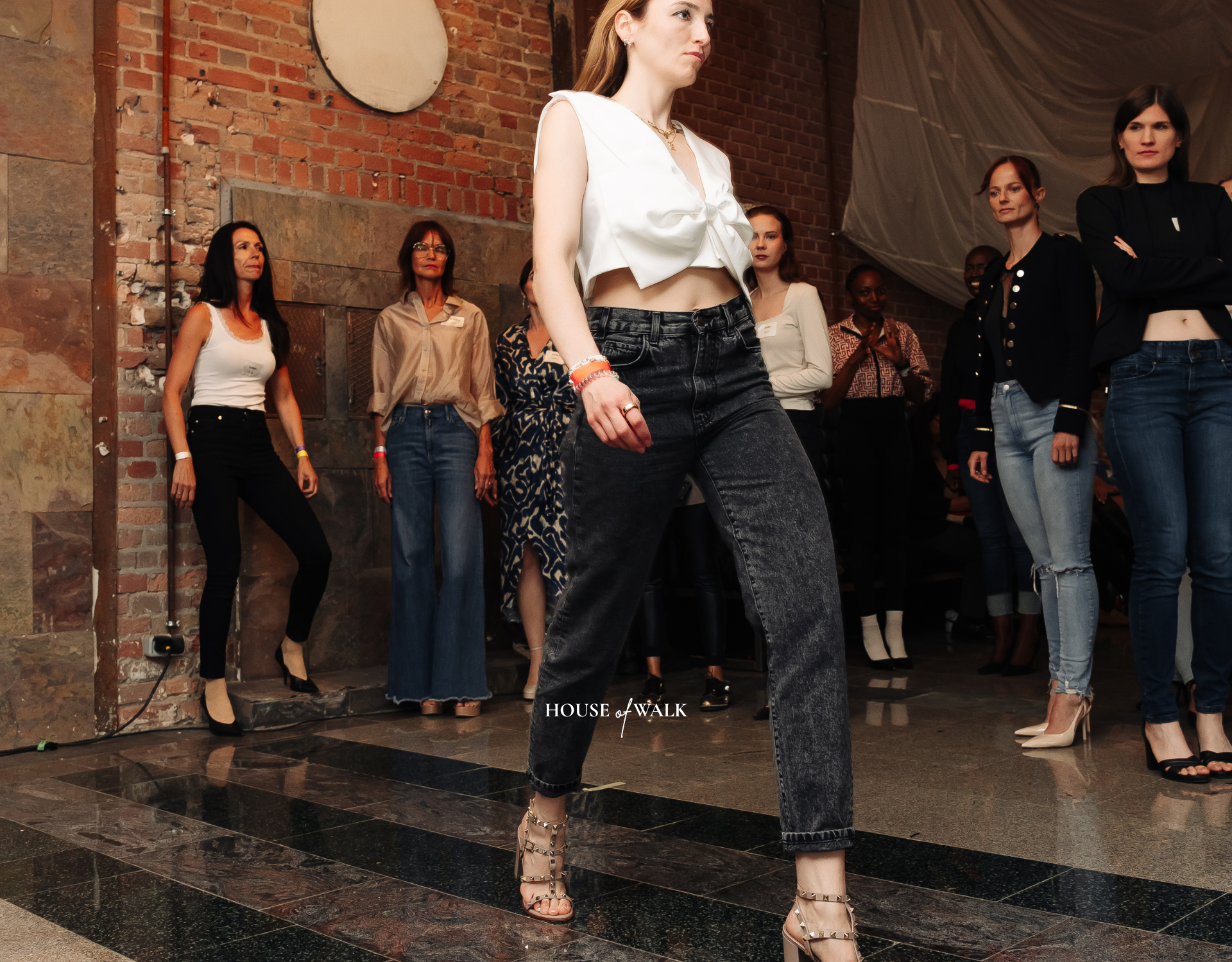 Frauen beim Mode-Event, Frau in der Vordergrund trägt schwarzer Jeans, weiße Top mit Schleife, und High Heels, im Hintergrund stehen weitere Frauen in unterschiedlichen Outfits, die Wände sind aus Backstein, der Boden ist aus Fliesen.