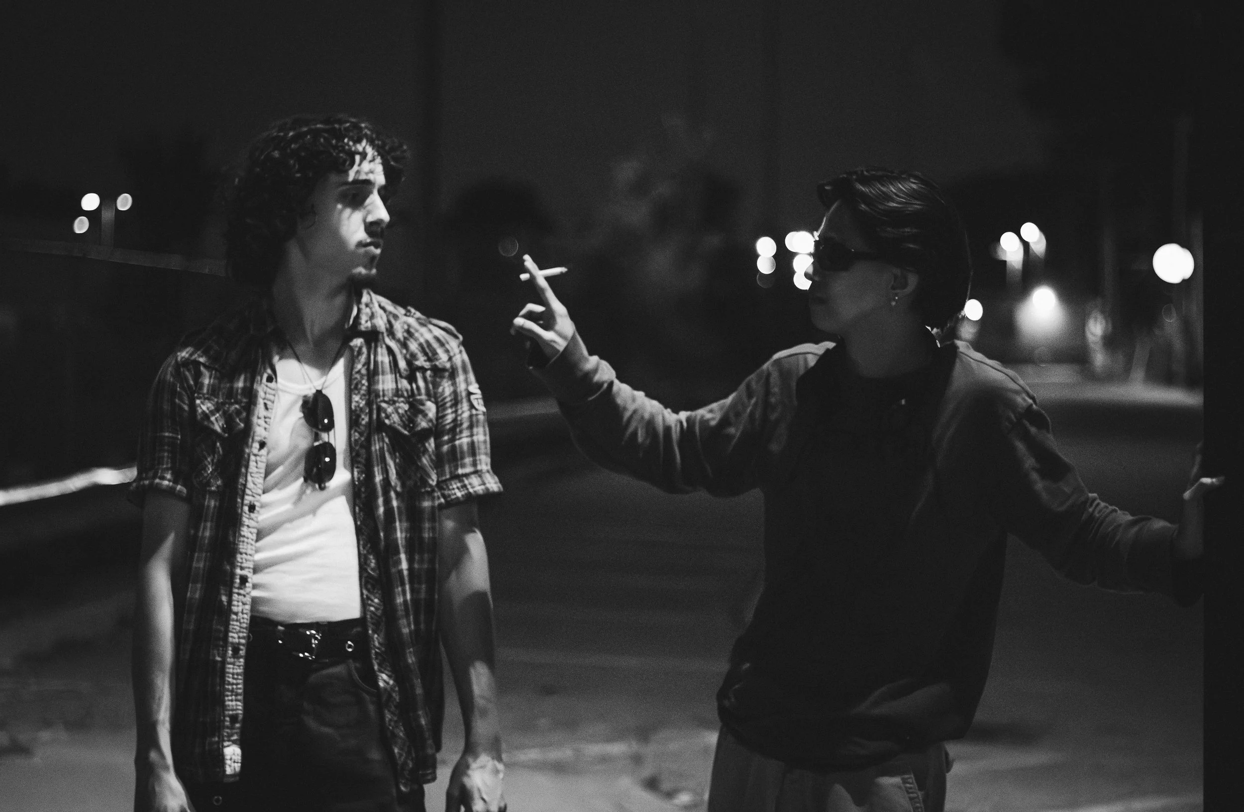 Two young men stand on a street at night, one smoking and the other gesturing with his hands. Both are dressed casually, one with sunglasses and the other with a plaid shirt, in black and white.