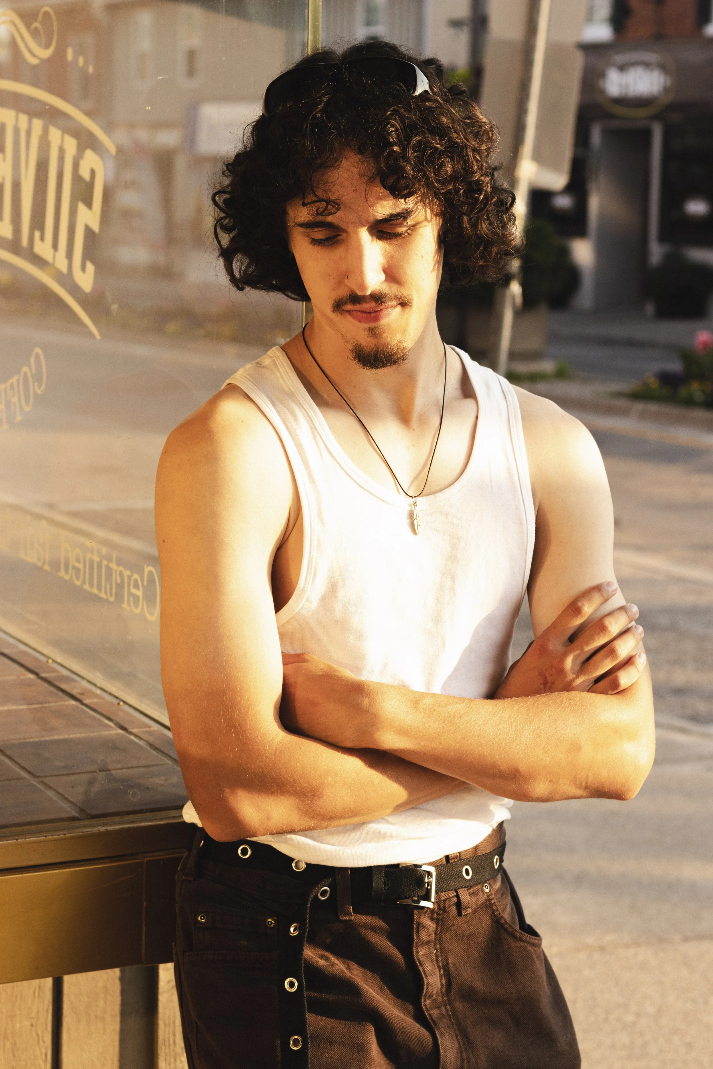A young man with curly hair and a goatee, wearing a white tank top, necklace, and dark pants, leaning against a glass window on a city street at sunset.