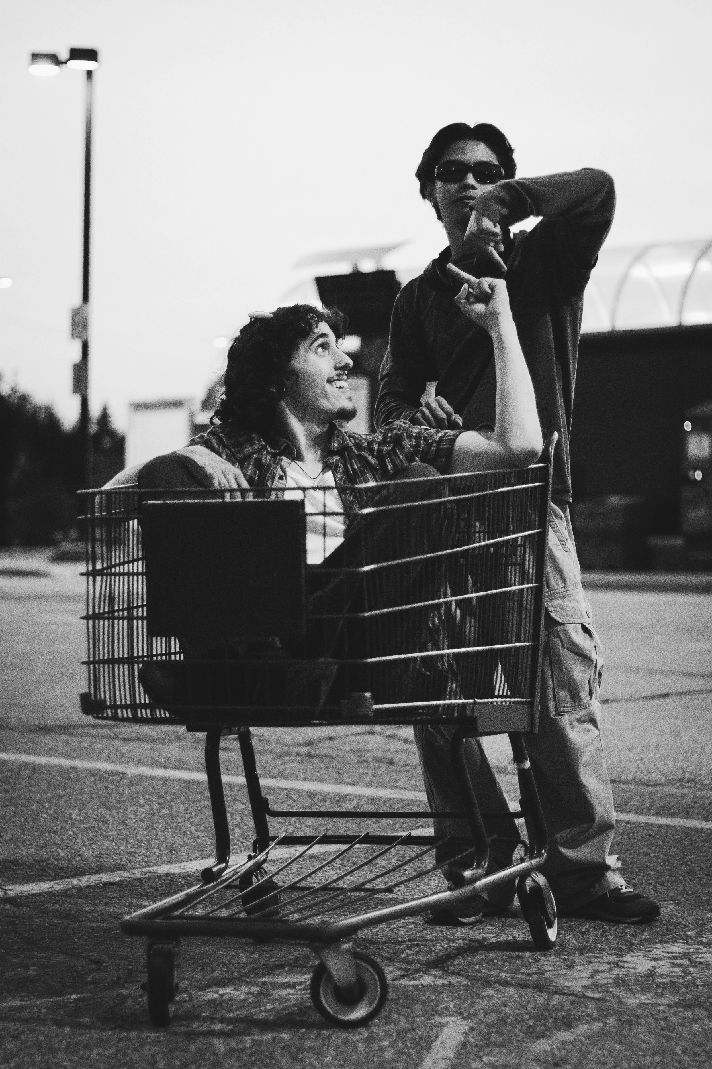Two young men, one sitting in a shopping cart and the other standing next to him, are having fun outdoors in an urban setting during dusk, with streetlights in the background.