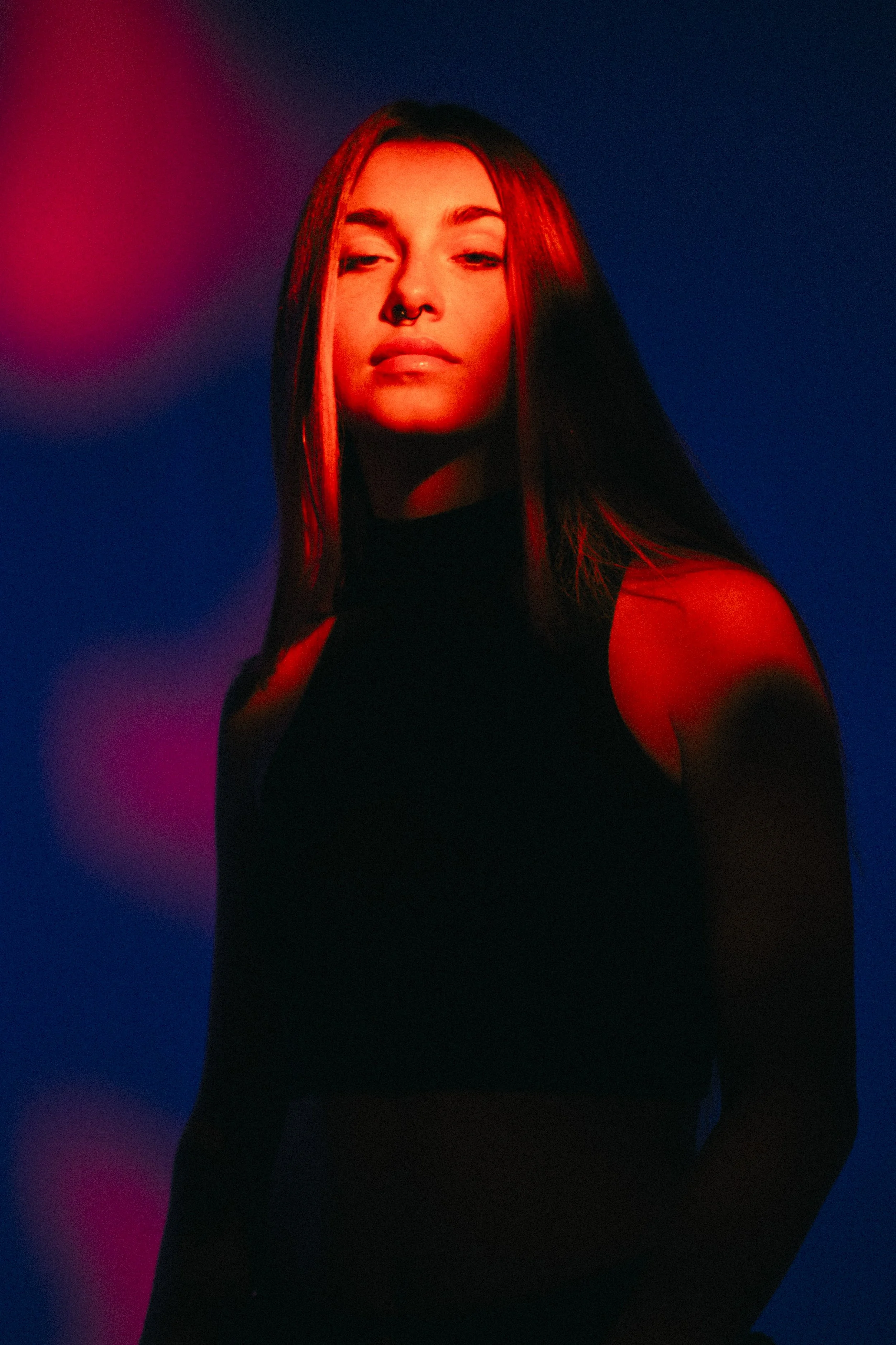 A woman with long dark hair, wearing a black sleeveless top, is illuminated by red and blue lighting against a dark background.