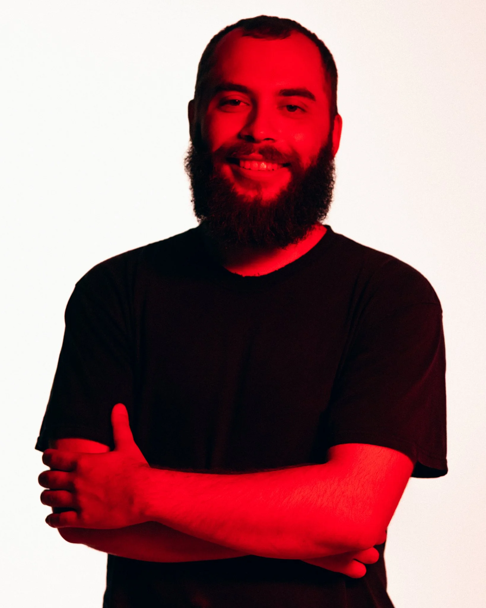 A man with a beard is smiling, crossed arms, wearing a black t-shirt, illuminated with red light.