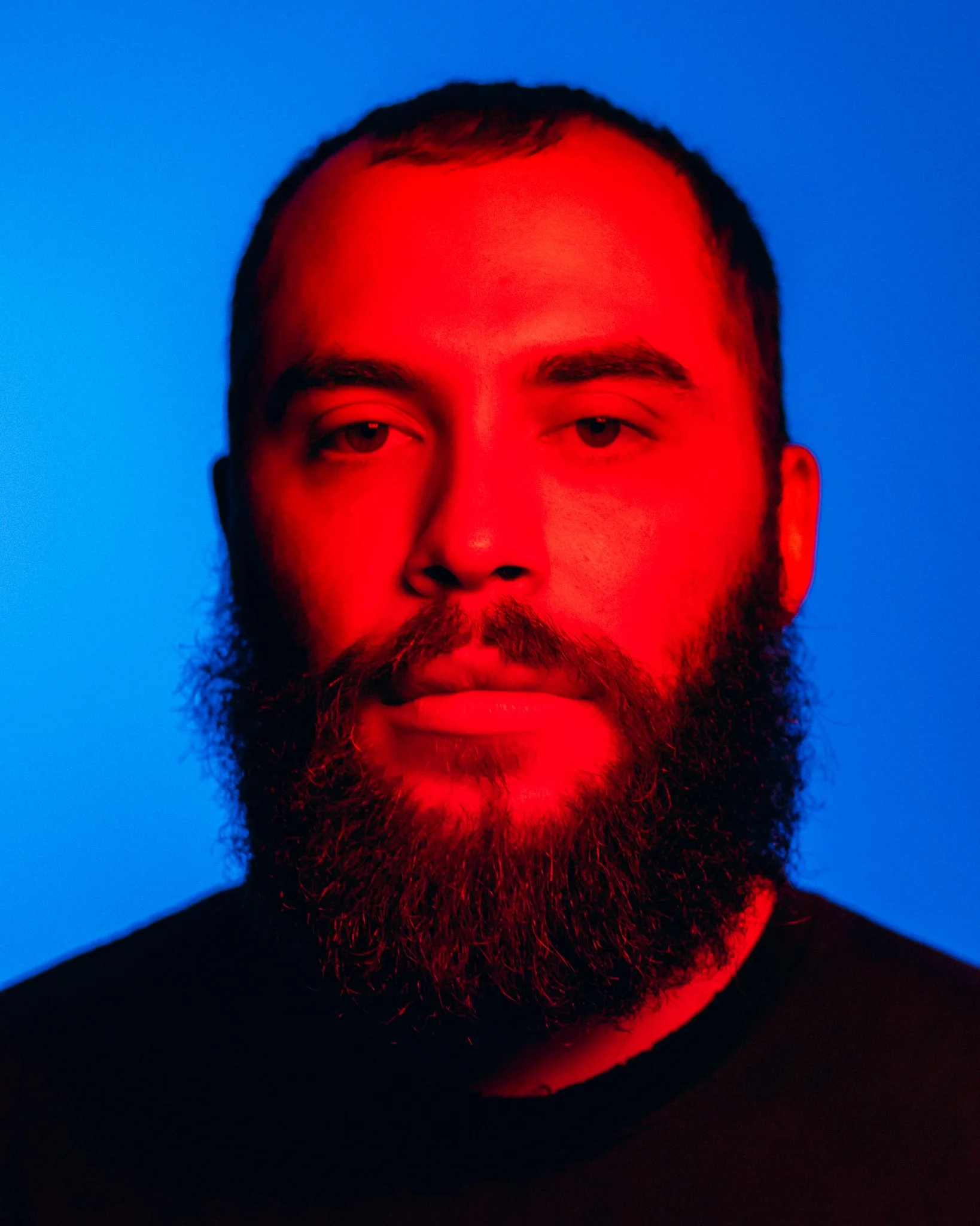 A portrait of a man with a full beard and short dark hair, illuminated with red lighting, against a blue background.