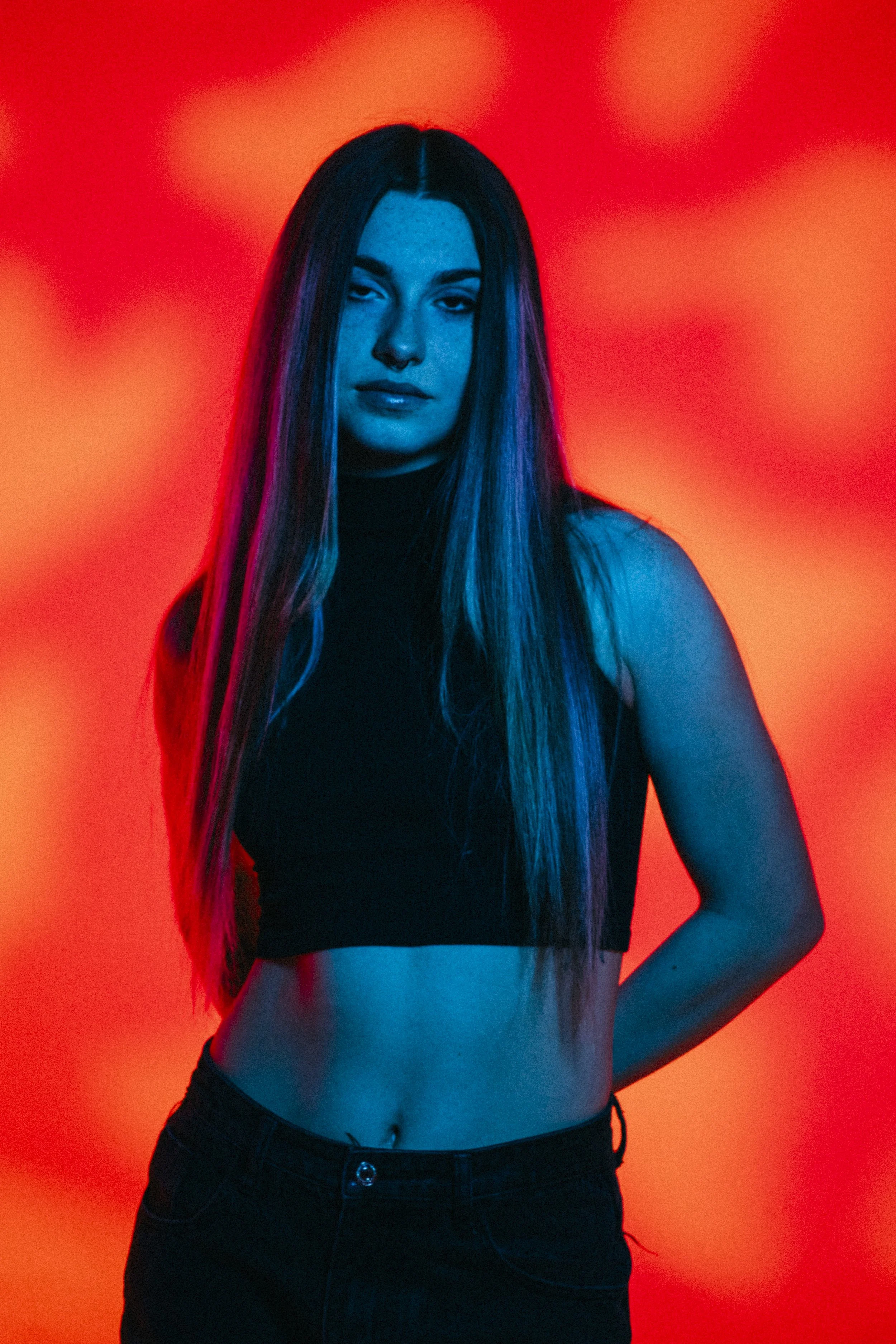 A young woman with long hair stands against a red and orange background, wearing a black sleeveless crop top and black pants.