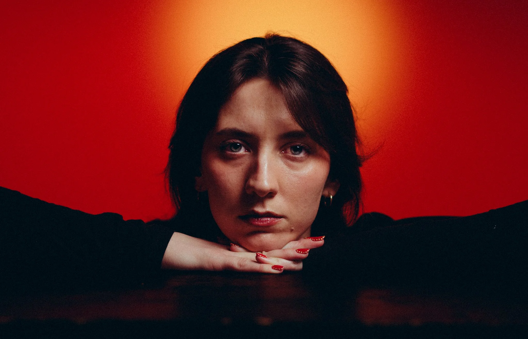 Portrait of a woman with dark hair, resting her chin on her hands, against a warm orange background.