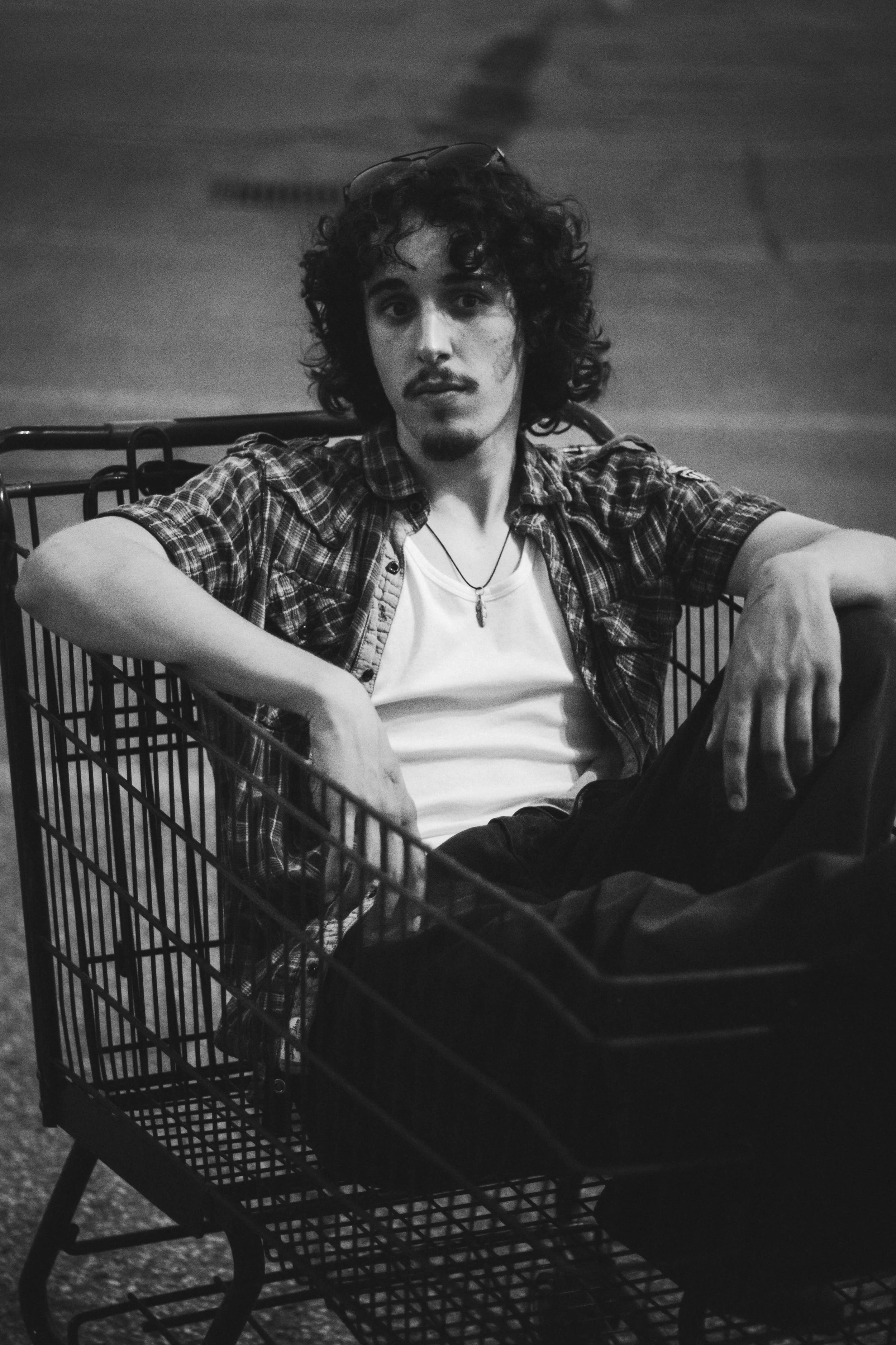 A young man with curly hair and a mustache sits in a shopping cart, looking at the camera. He's wearing a plaid shirt over a white t-shirt and has a necklace. The photo is black and white.