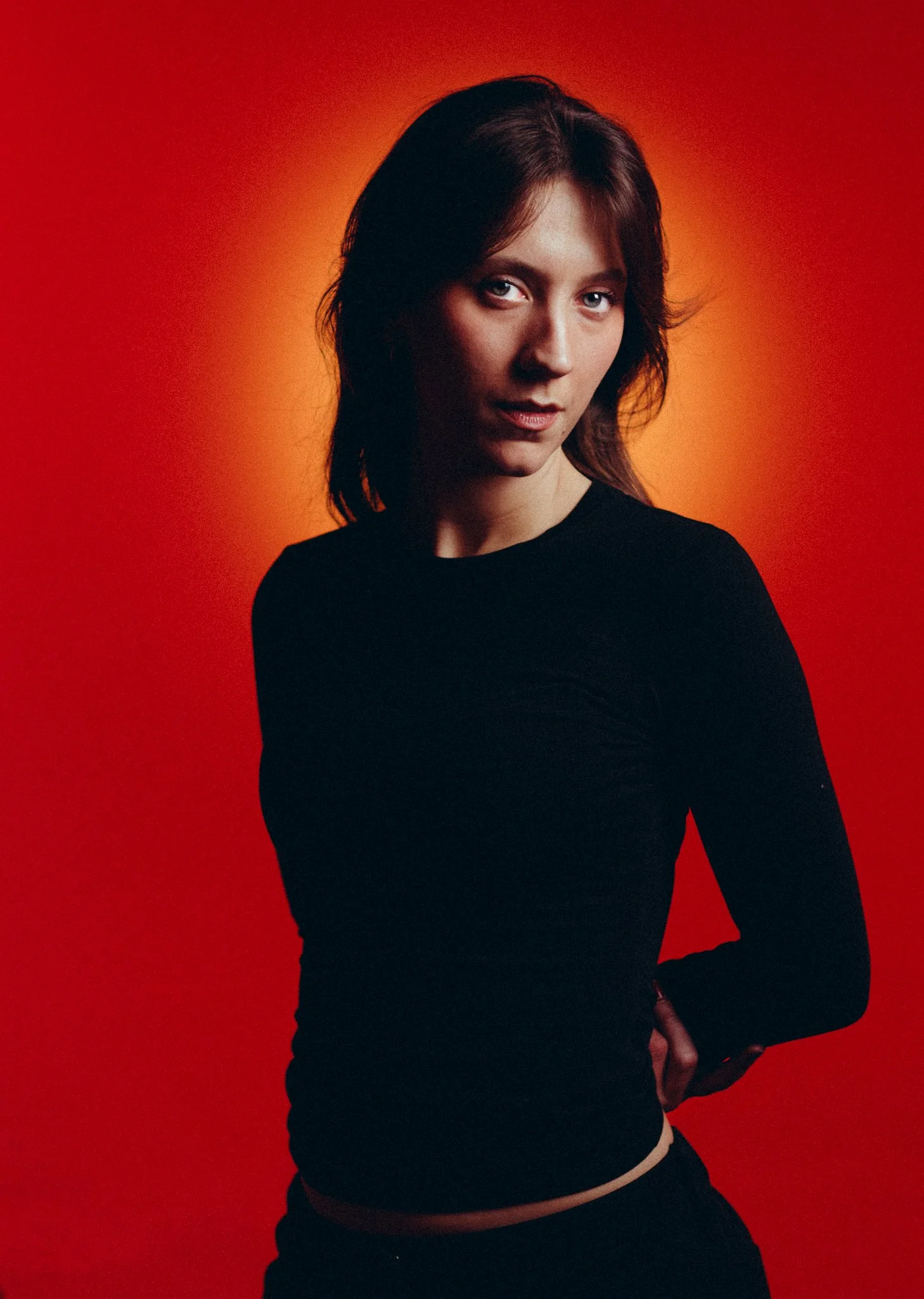 A woman with reddish-brown hair in a black long-sleeve top posing against a red-orange gradient background.