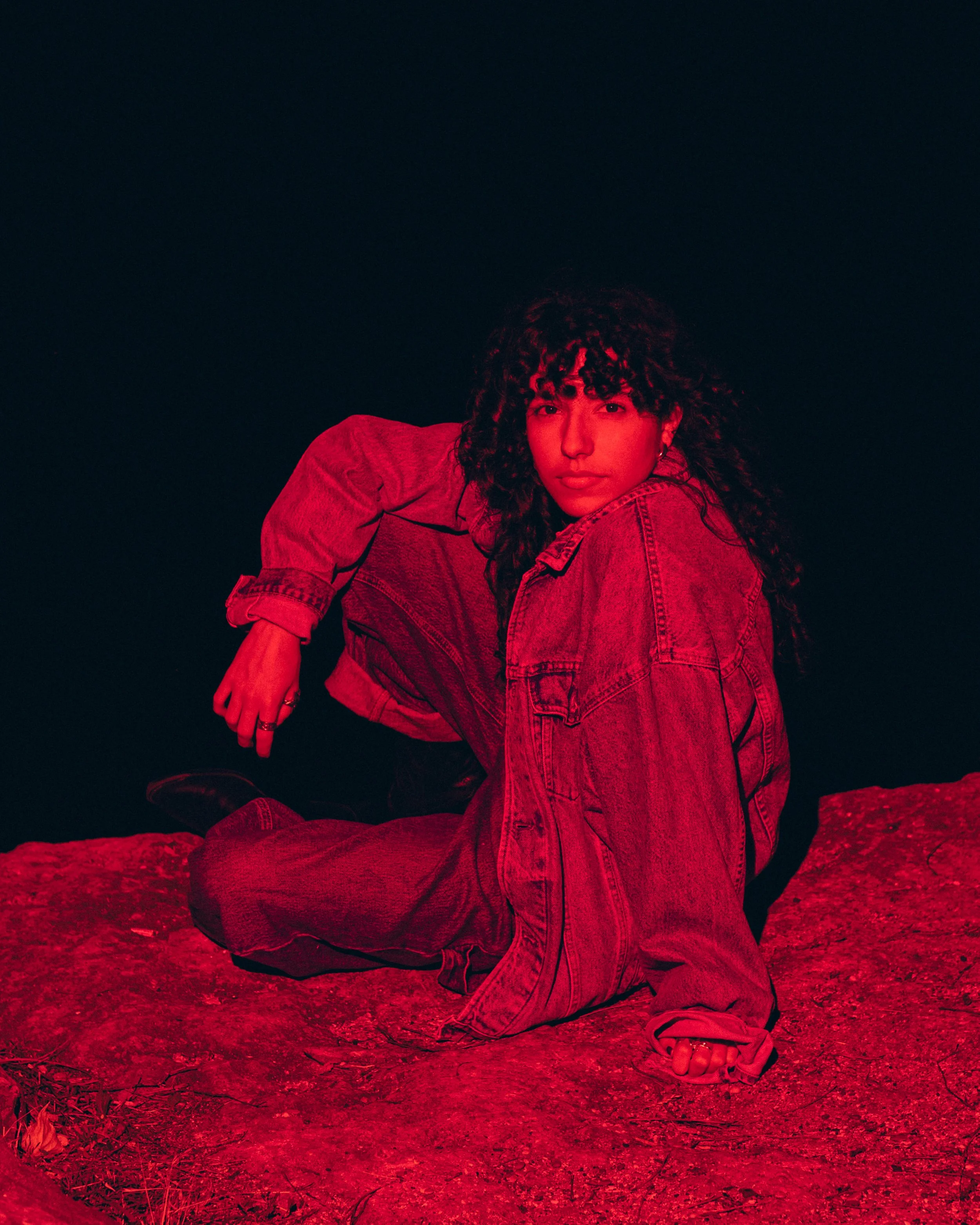 Person with curly hair wearing a denim jacket and pants, sitting on a large rock at night with red lighting.