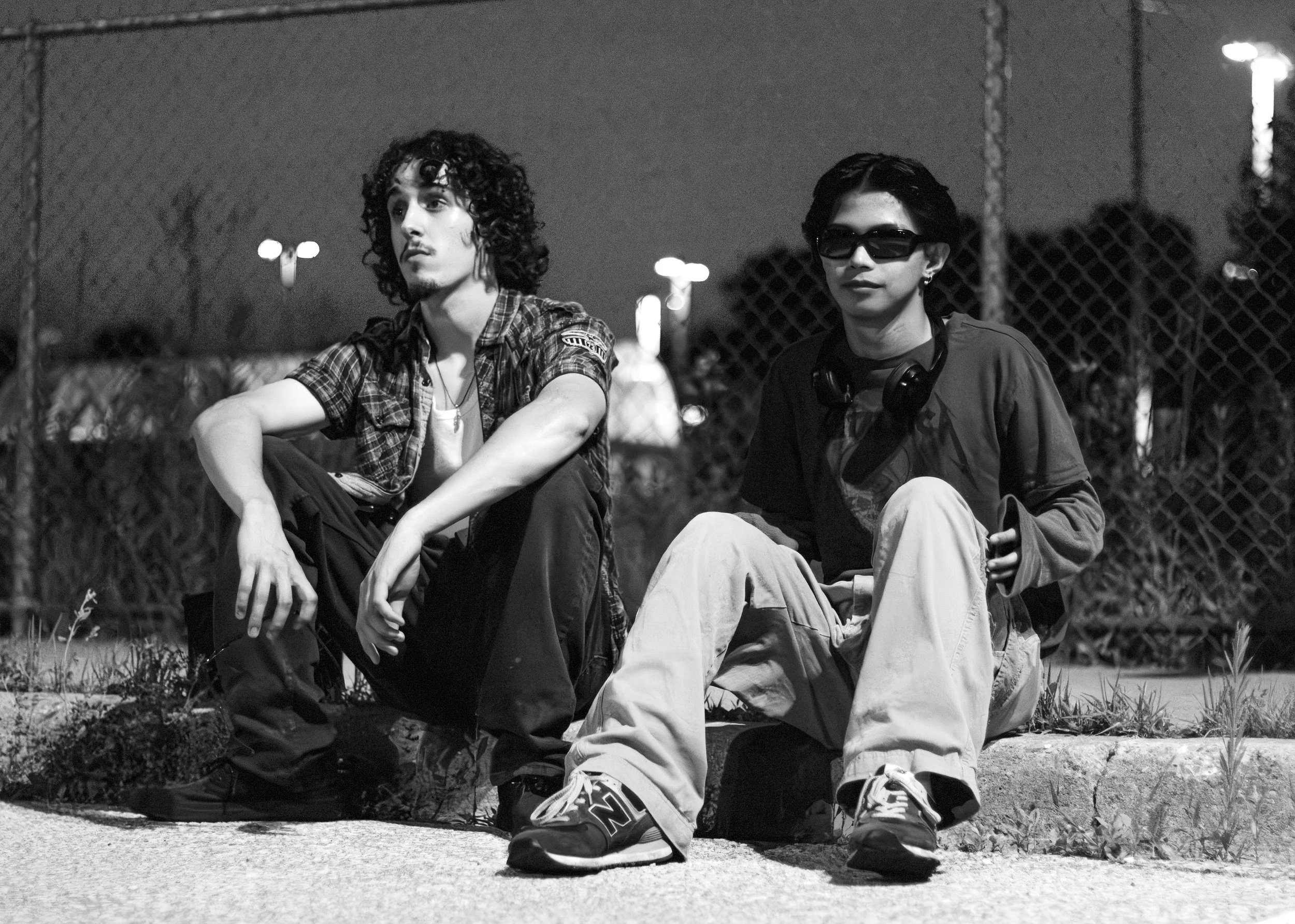 Two young men sitting on a curb at night, near a chain-link fence with city lights in the background. One has curly hair, wears a plaid shirt, and has headphones around his neck. The other wears sunglasses, a dark jacket, and light-colored pants.