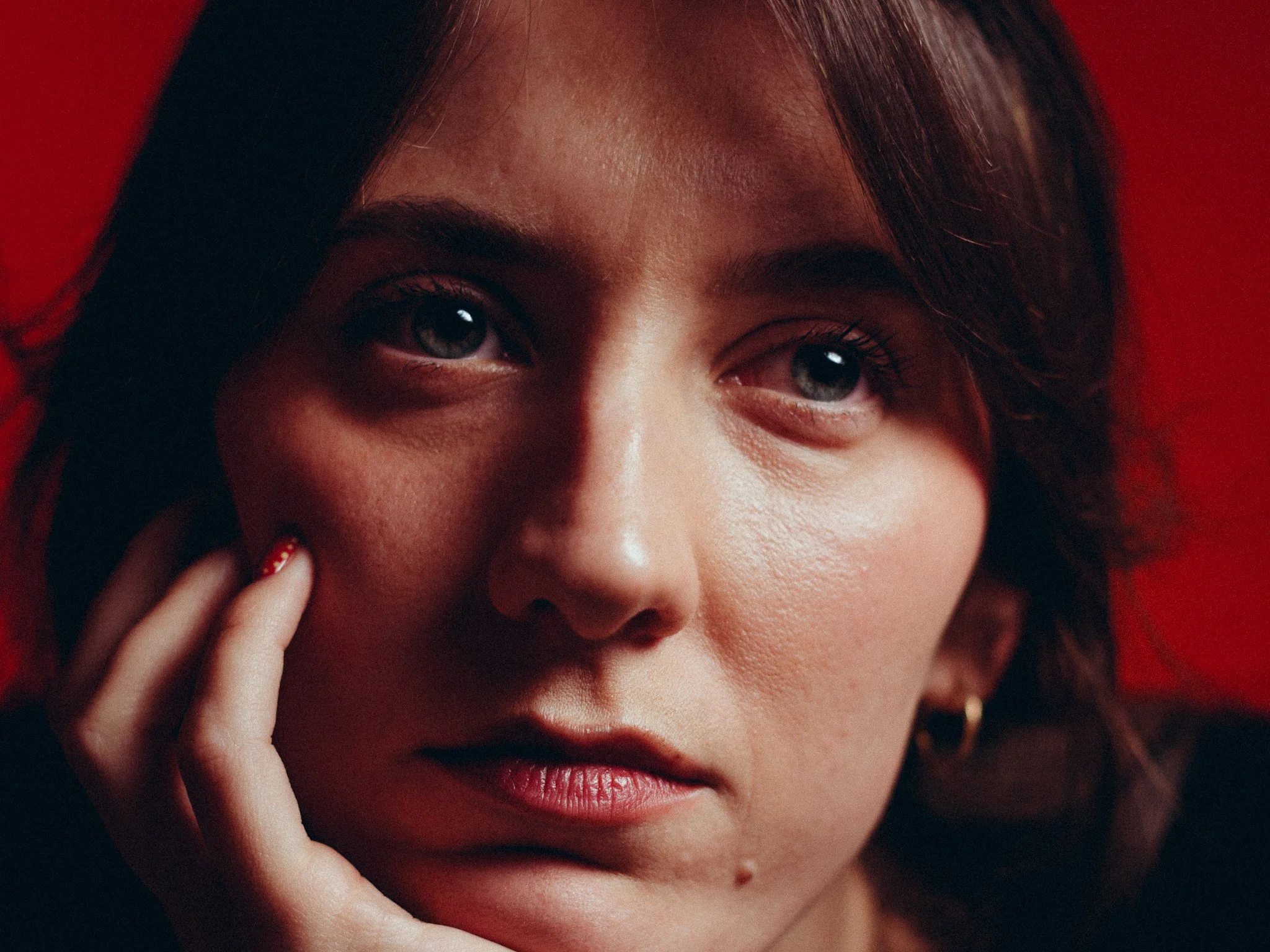 A close-up of a woman with short, wavy brown hair and blue eyes, resting her face on her hand against a red background.