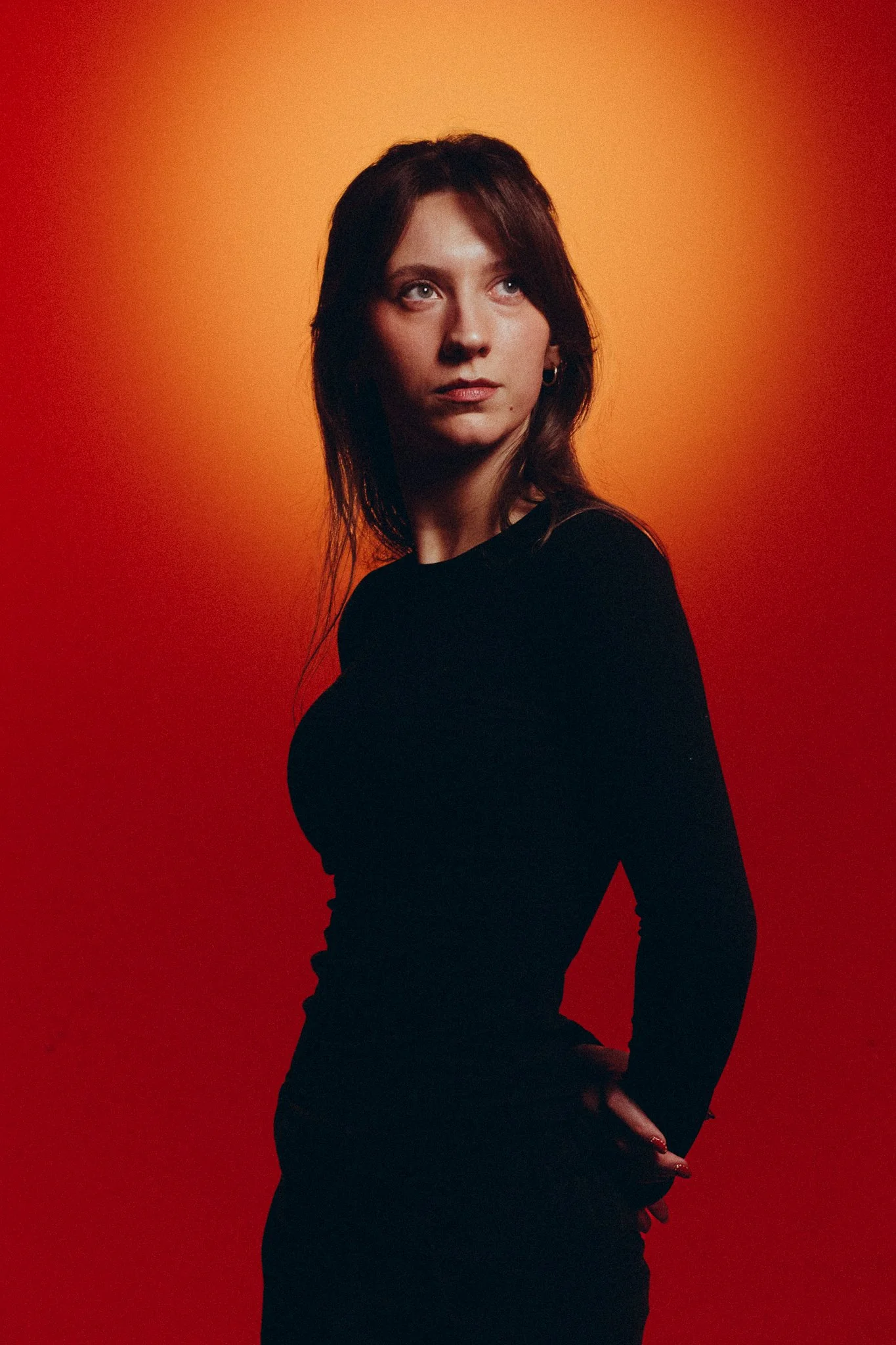 Portrait of a woman with long brown hair wearing a black top, standing against a gradient background of orange and red.
