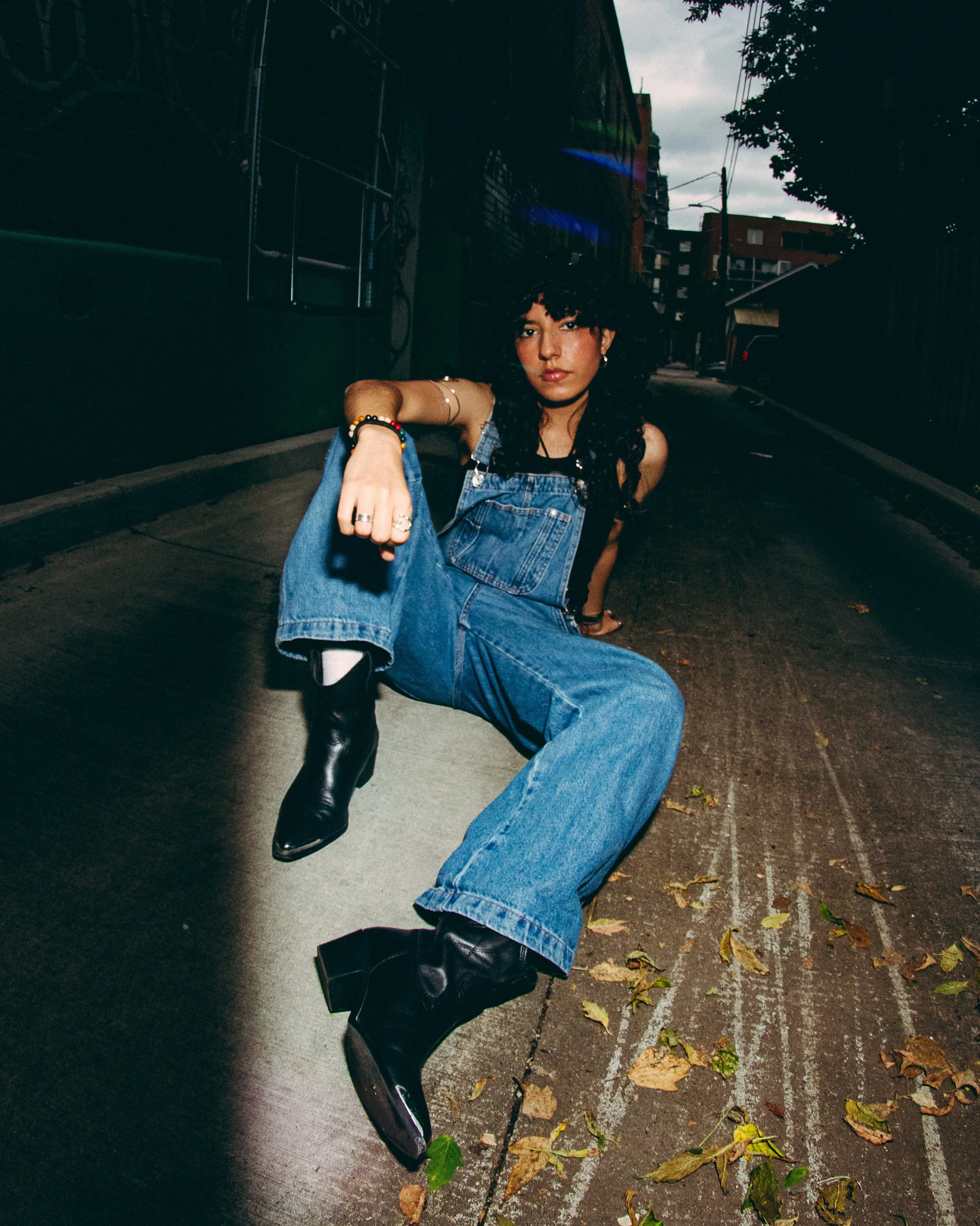 A woman with curly black hair poses on an urban street at night, sitting on the pavement with one knee raised. She wears blue jeans, black heeled boots, a black top, and layered jewelry, with a confident expression, dark makeup, and accessories like 