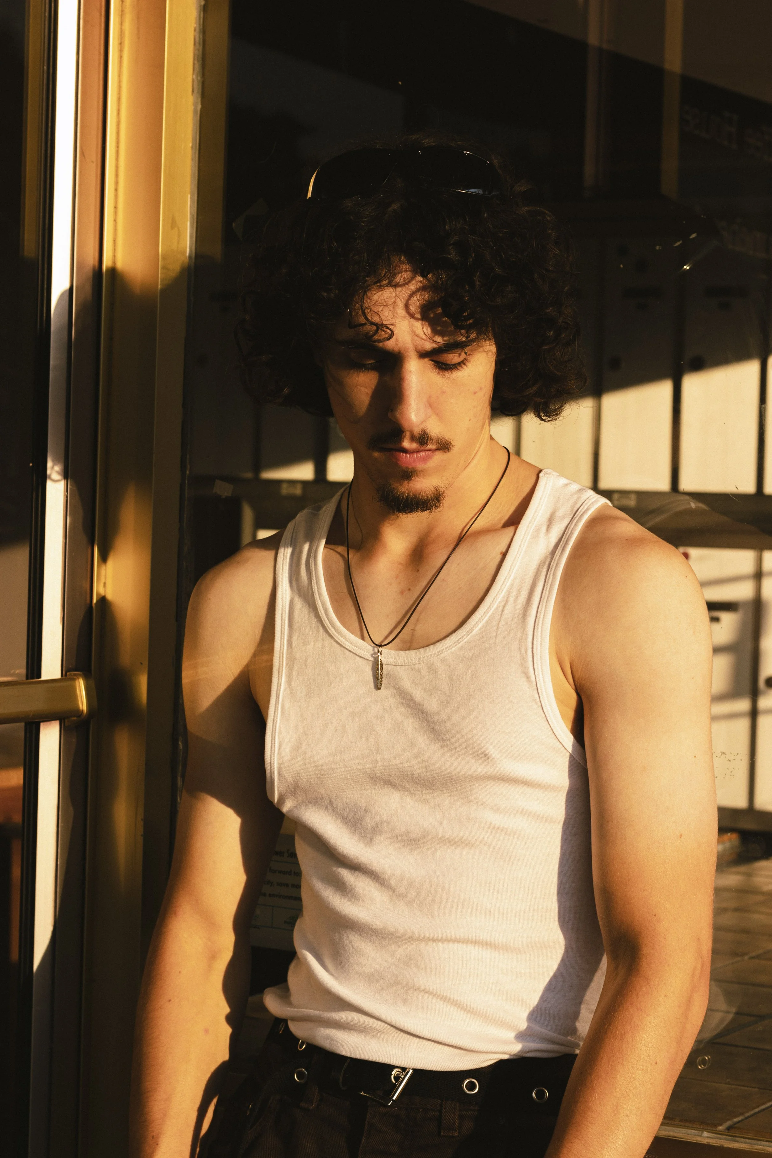 A young man with curly dark hair, a mustache, and a goatee, wearing a white tank top and sunglasses on his head, standing outside next to a glass door, looking down with a shadow cast across his face from the sunlight.